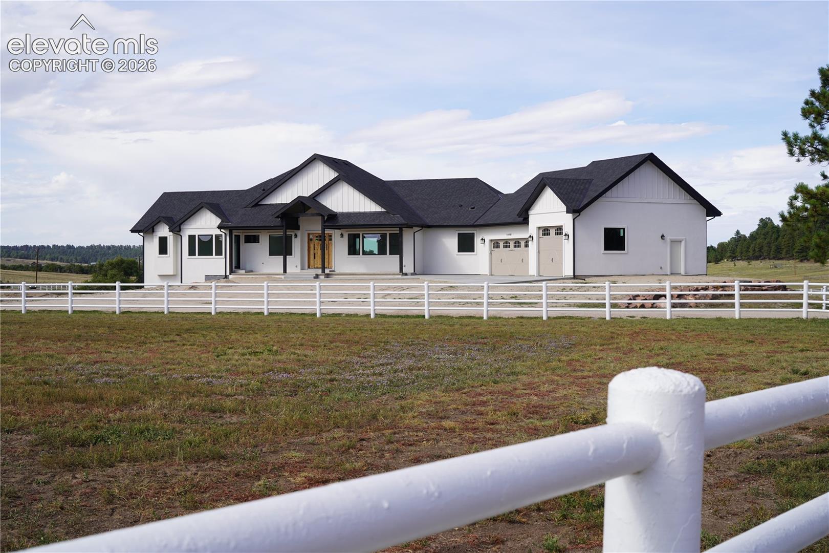 White exterior with black trim, black shingle roof, attached two-car garage, and a white fence