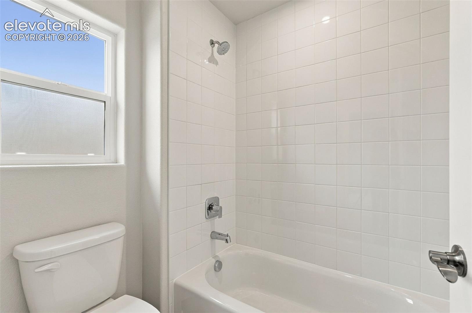 Full-height white square tile tub surround, integrated soaking tub, chrome finish plumbing fixtures, and single-pane window with frosted glass