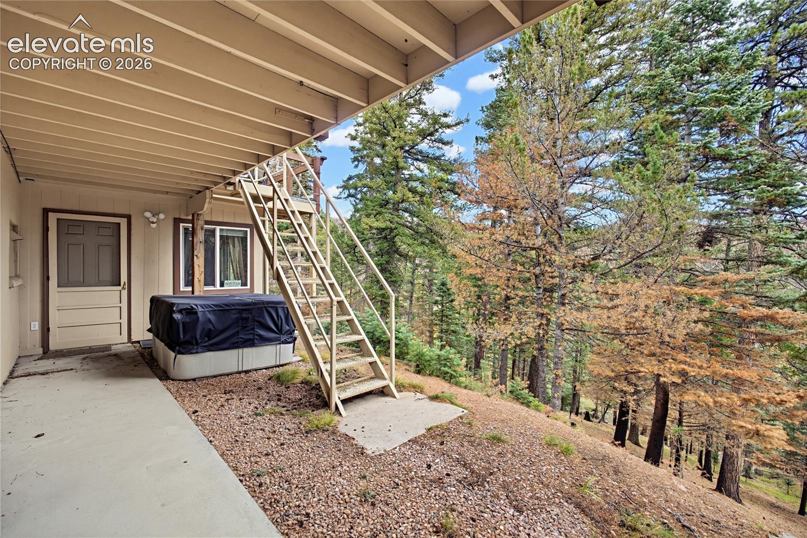 Covered concrete patio featuring external metal staircase, exterior entry door, and hillside lot with mature evergreen trees