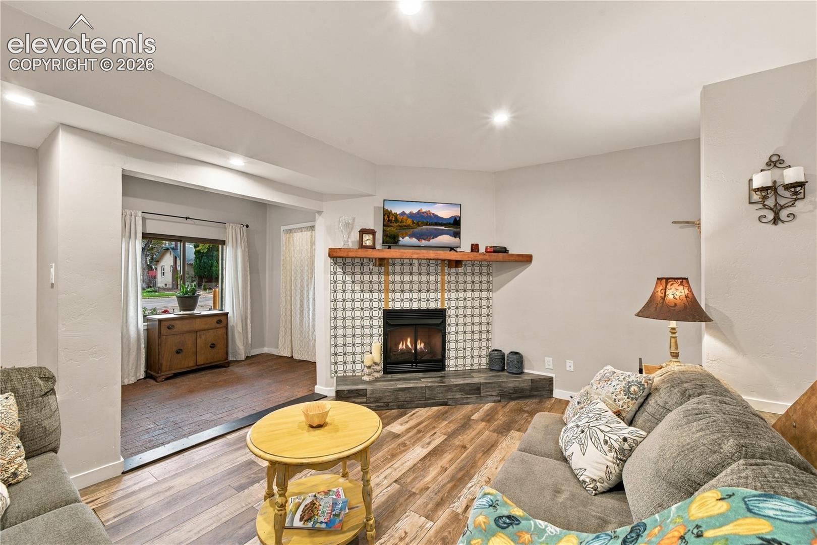 Fireplace featuring a patterned tile surround, stone-finish hearth, and wood mantel, complemented by wood-finish flooring and recessed lighting