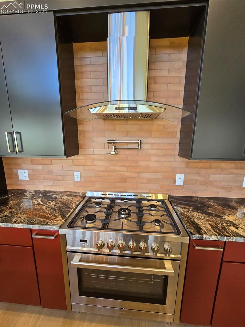 Kitchen featuring dark stone countertops, exhaust hood, decorative backsplash, and stainless steel range oven
