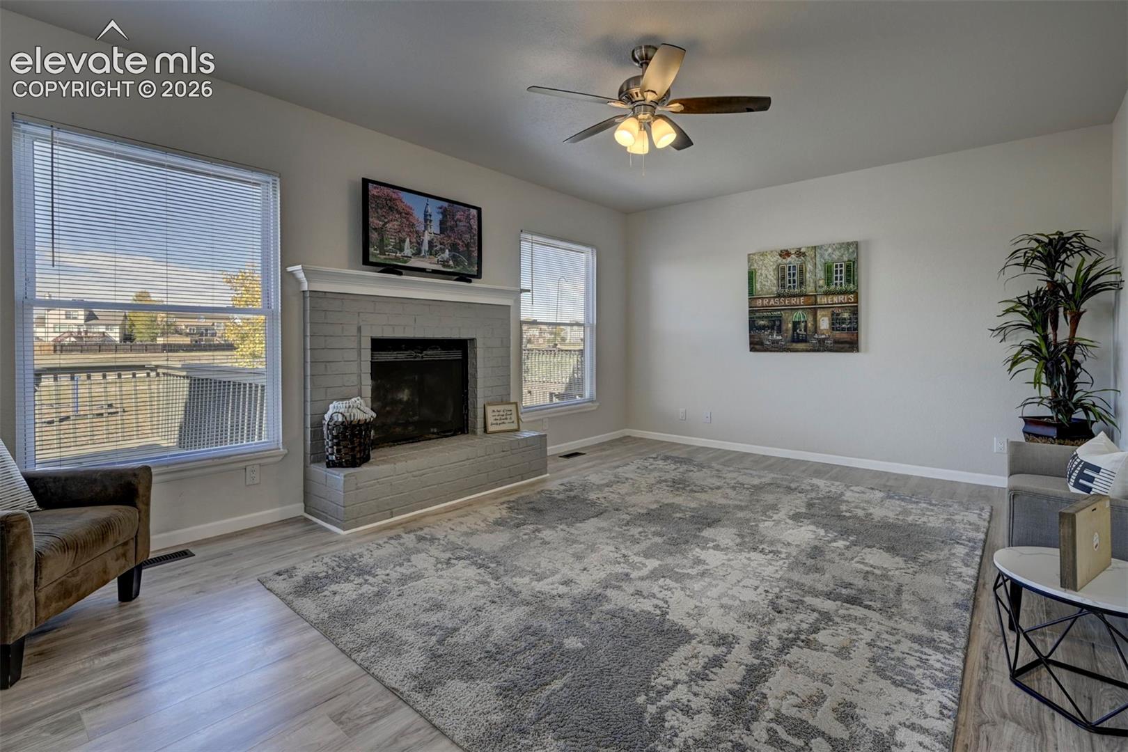 Painted brick fireplace with mantel, wood-finish flooring, ceiling fan with integrated lighting, and dual windows