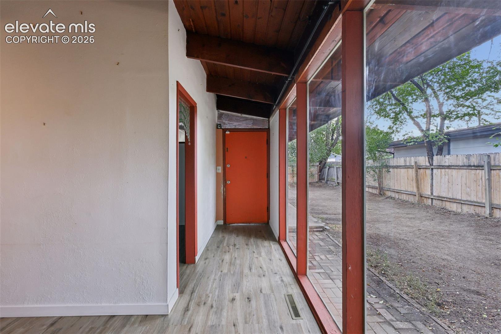 Wood-finish flooring with a vibrant orange entry door, exposed wood ceiling beams, and a full wall of glass panels