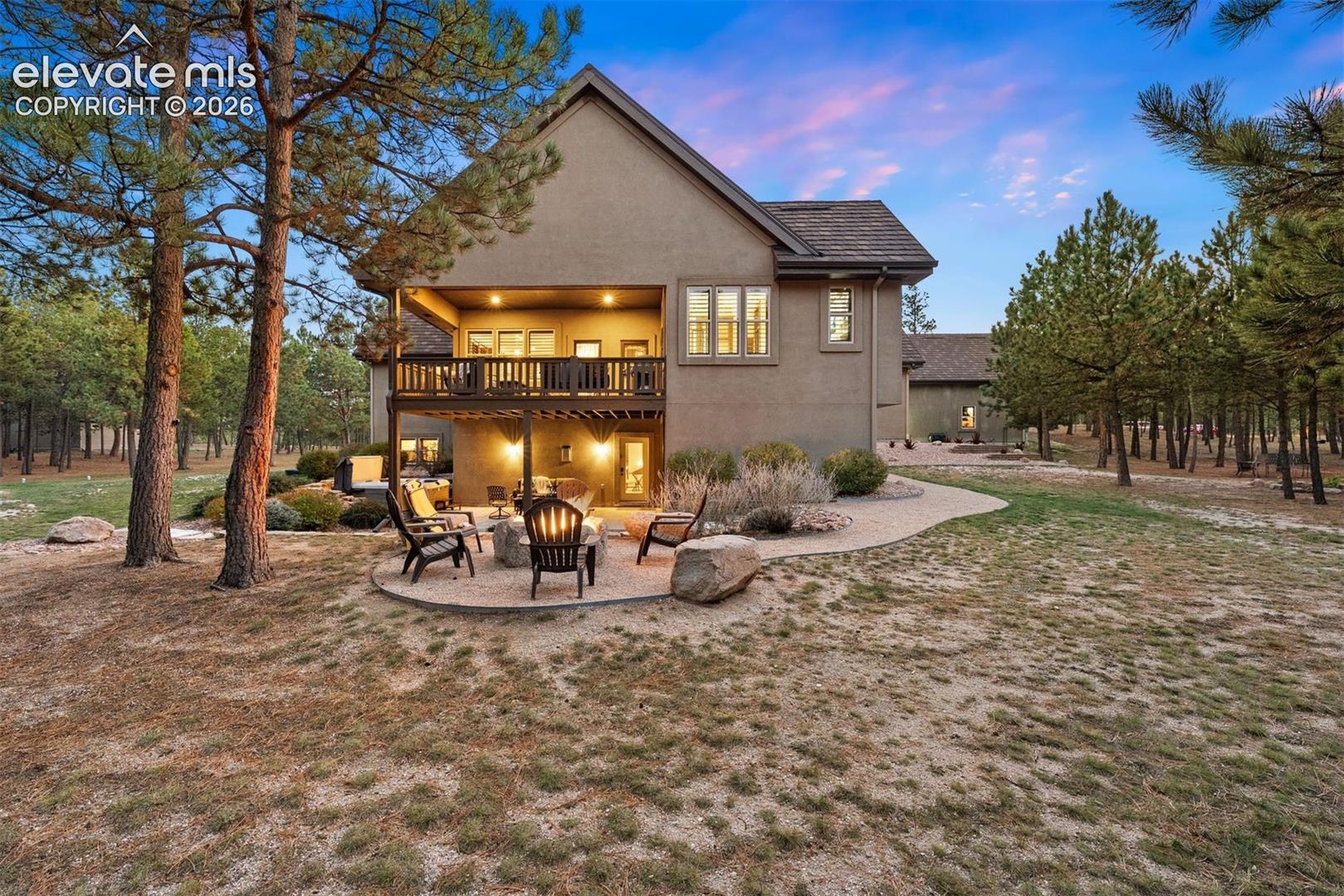 Stucco exterior residence featuring a covered upper-level deck, ground-level stone-paver patio with a built-in stone fire pit, and expansive grounds with mature pine trees