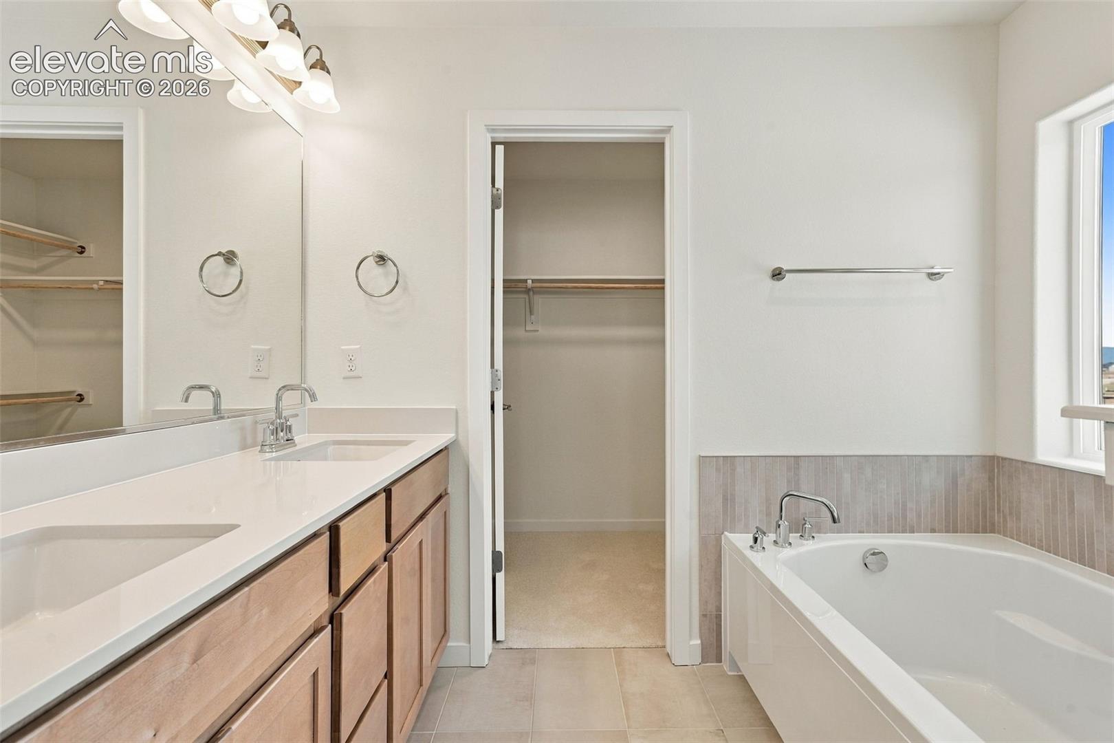 Dual vanity with integrated sinks, light wood-finish cabinetry, built-in soaking tub, and tile flooring