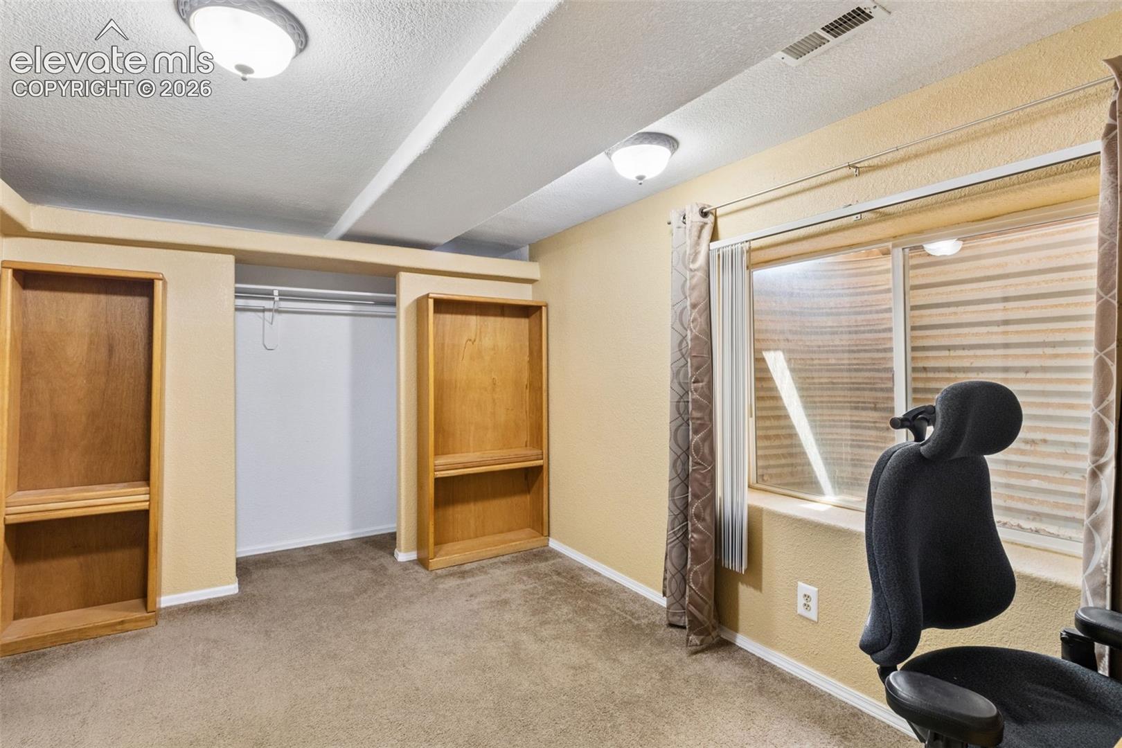 Room featuring two built-in wood-finish shelving units, a recessed closet with an overhead rod, dual flush-mount ceiling light fixtures, and a recessed window with an egress-style well cover