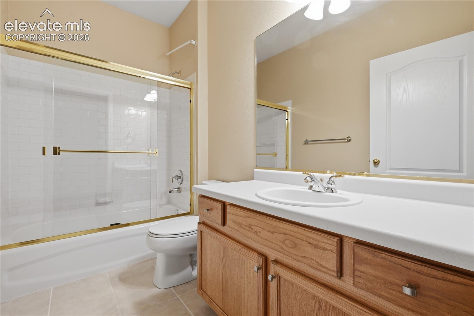 Bathroom featuring a vanity with wood-finish cabinetry, a solid surface countertop with an integrated basin, a large wall-mounted mirror, and a shower-tub combination with gold-finish frame and subway tile surround