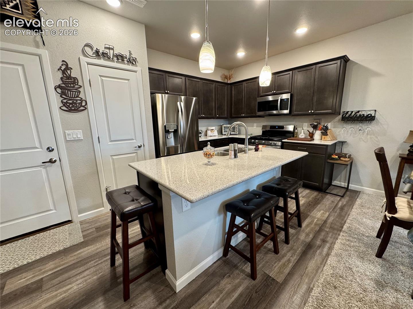 Kitchen featuring dark-toned cabinetry, light-colored stone-finish countertops, stainless steel appliances, a central island with an integrated sink, and pendant lighting