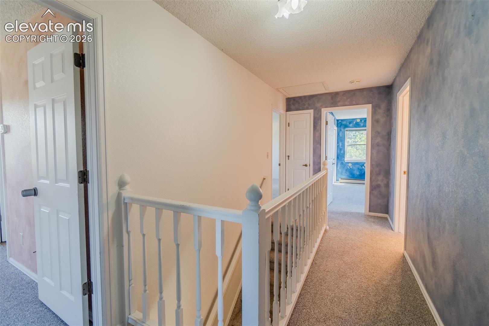 Carpeted hallway with white interior doors, a white banister, cool paint and attic access
