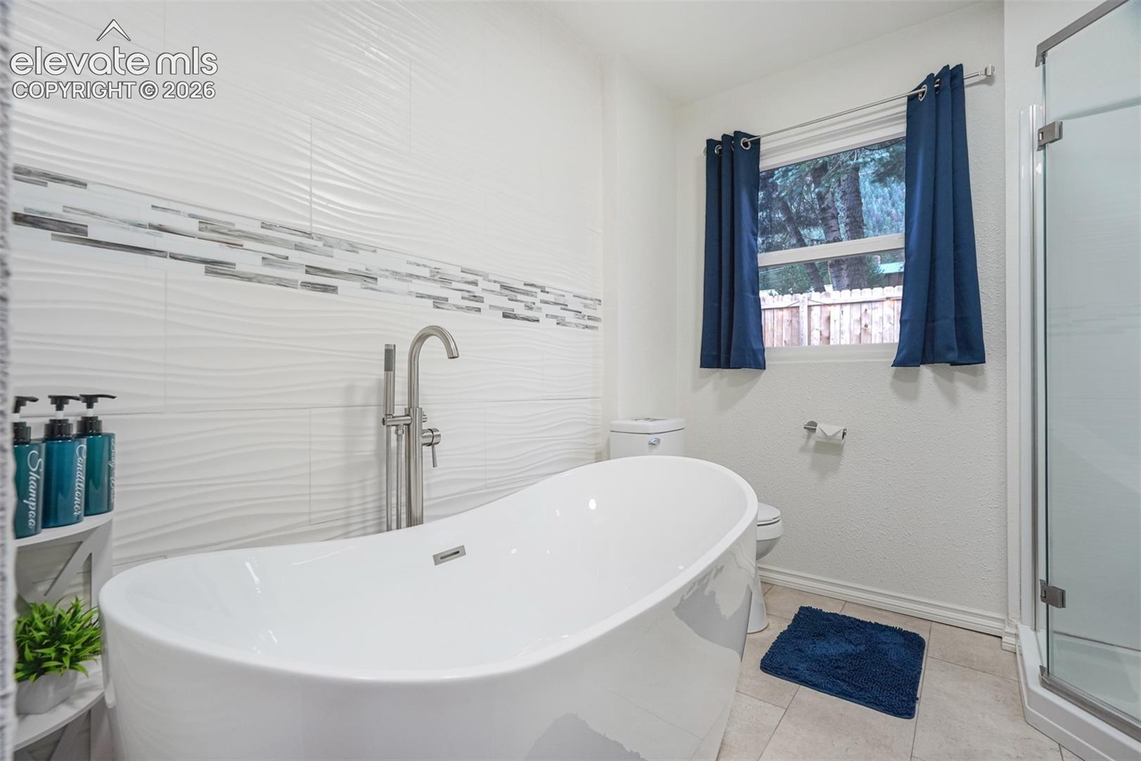 Freestanding soaking tub with floor-mounted faucet, textured tile wall with decorative mosaic accent, and glass-enclosed shower
