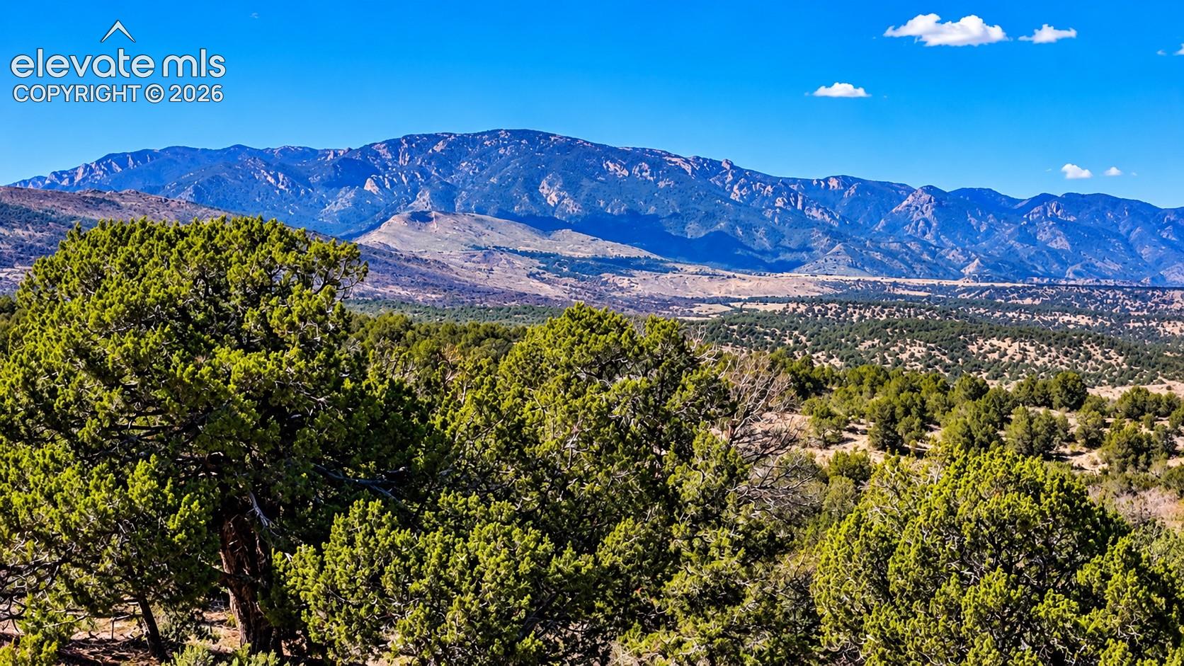 Breathtaking aerial panorama of rolling hills, scattered juniper trees, and wide-open ranchlands surrounded by distant mountain ranges.

