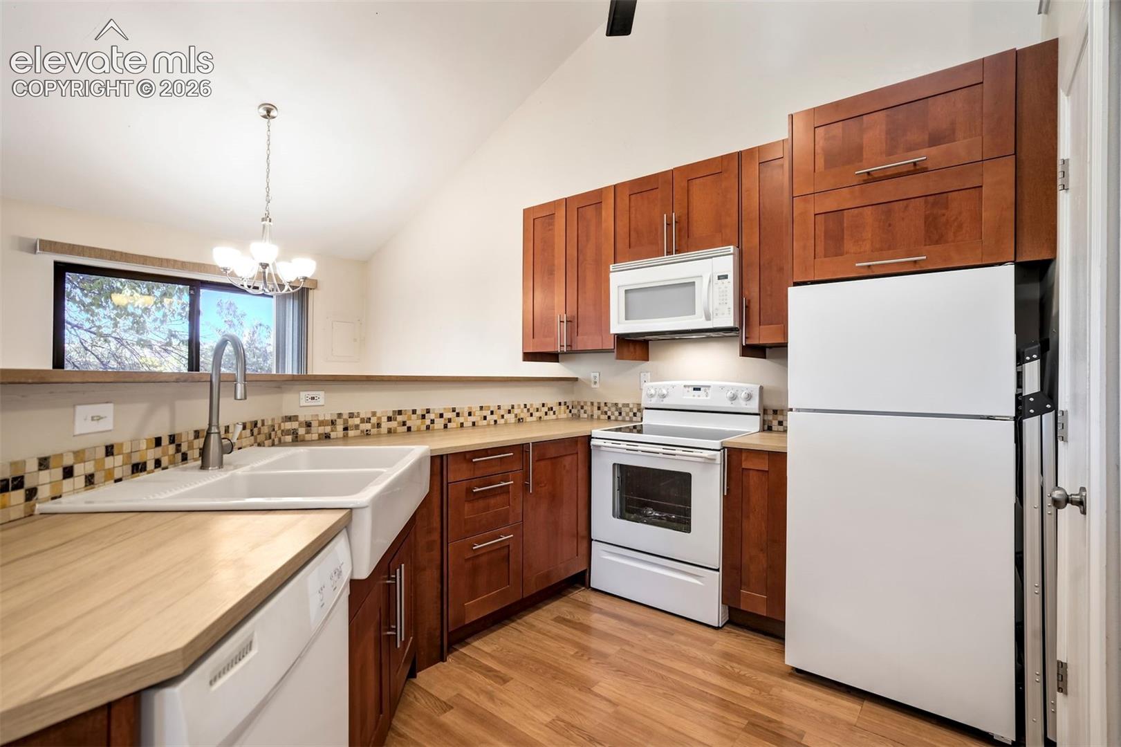 Kitchen featuring wood cabinetry, white farmhouse-style dual-basin sink with a gooseneck faucet, neutral countertops, tile backsplash, and vaulted ceilings