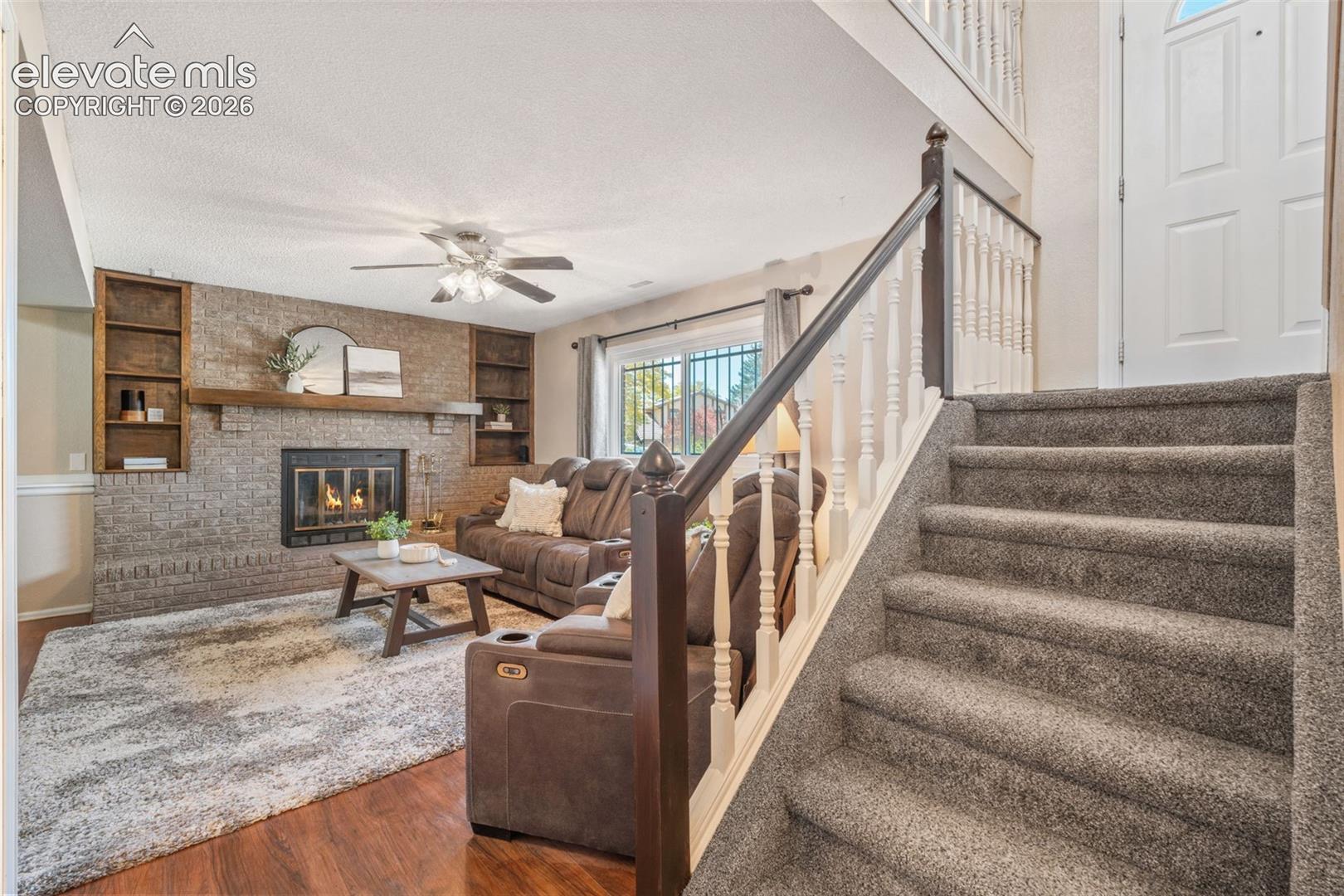 Living area featuring a brick fireplace with a wood mantel, built-in shelving, wood-finish flooring, and a staircase with a dark wood-finish railing and white balusters