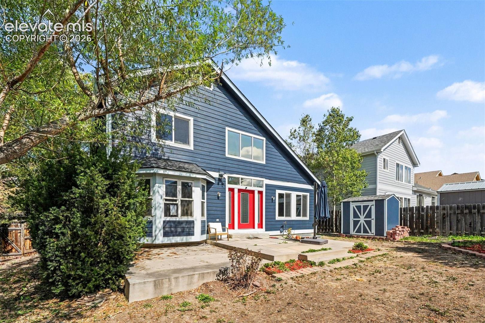Exterior featuring blue horizontal siding, white trim, red entry door, bay window with dark roof, and a paved patio area