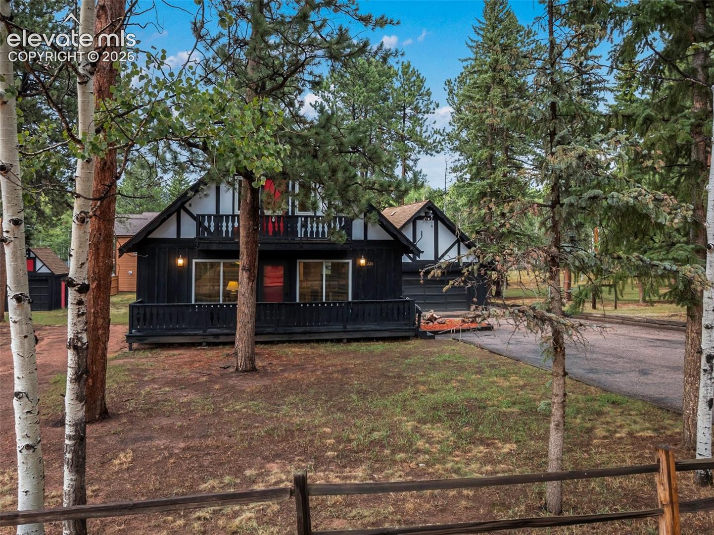 Black exterior with white trim, upper balcony, front porch, and gravel driveway, surrounded by mature trees