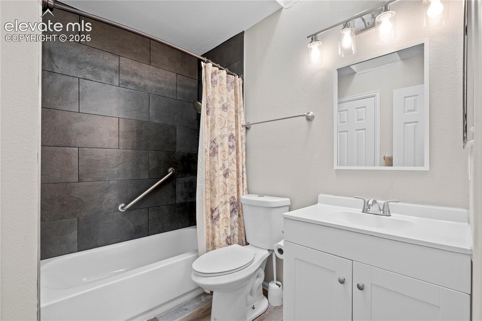 Bathroom featuring a white vanity cabinet with an integrated sink, polished metal faucet, wall-mounted mirror, four-light vanity fixture, and a tub-shower combination with dark large-format tile surround
