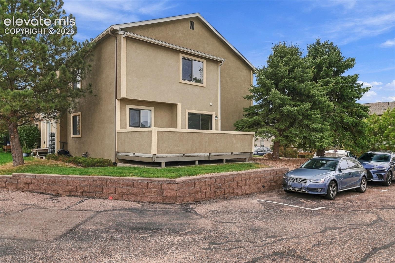 Multi-level stucco exterior featuring a raised wooden deck, decorative stone retaining wall, and mature coniferous landscaping