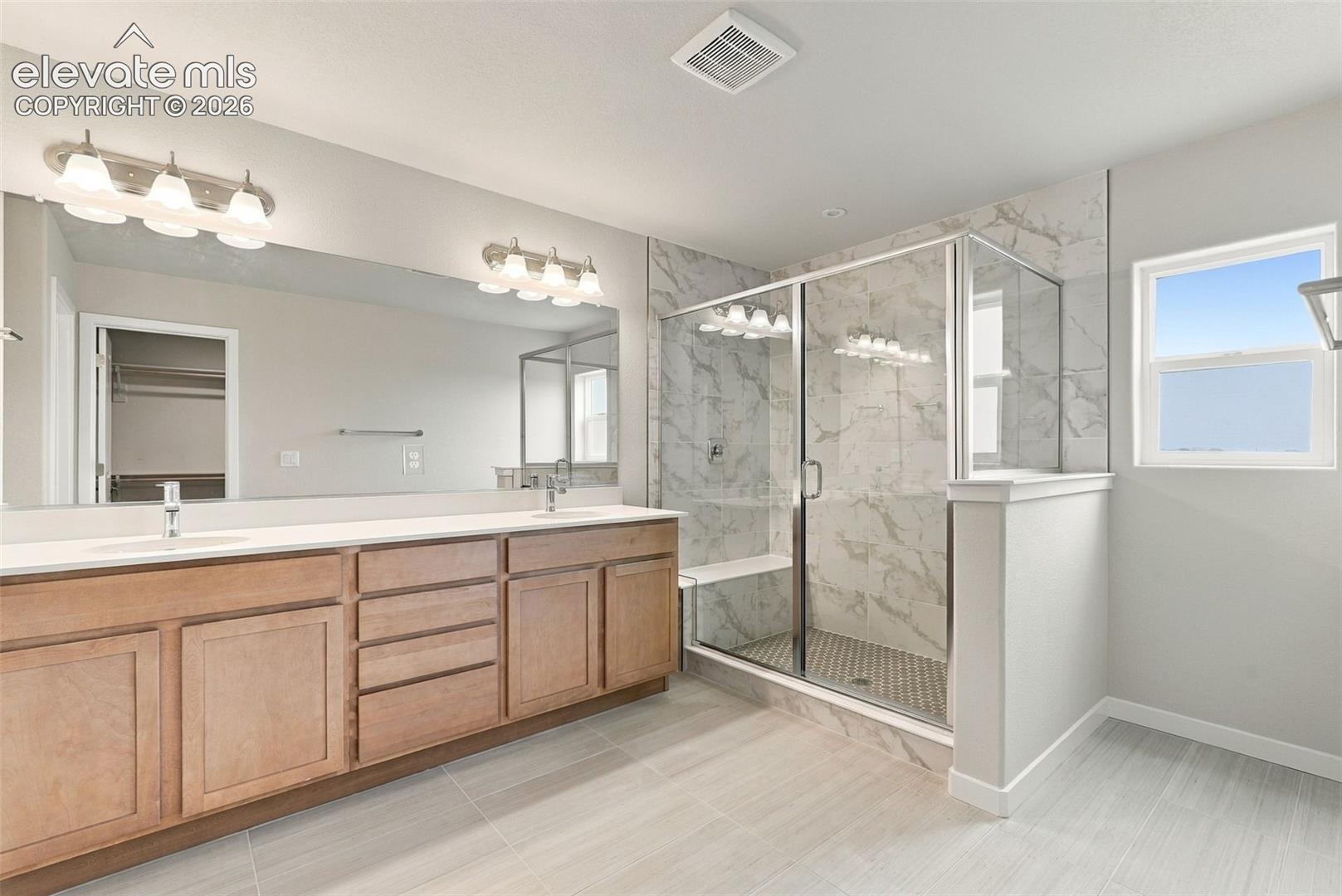 Double vanity with integrated sinks, wood-finish cabinetry, expansive wall-mounted mirror, walk-in shower with stone-patterned tile surround, and tile flooring