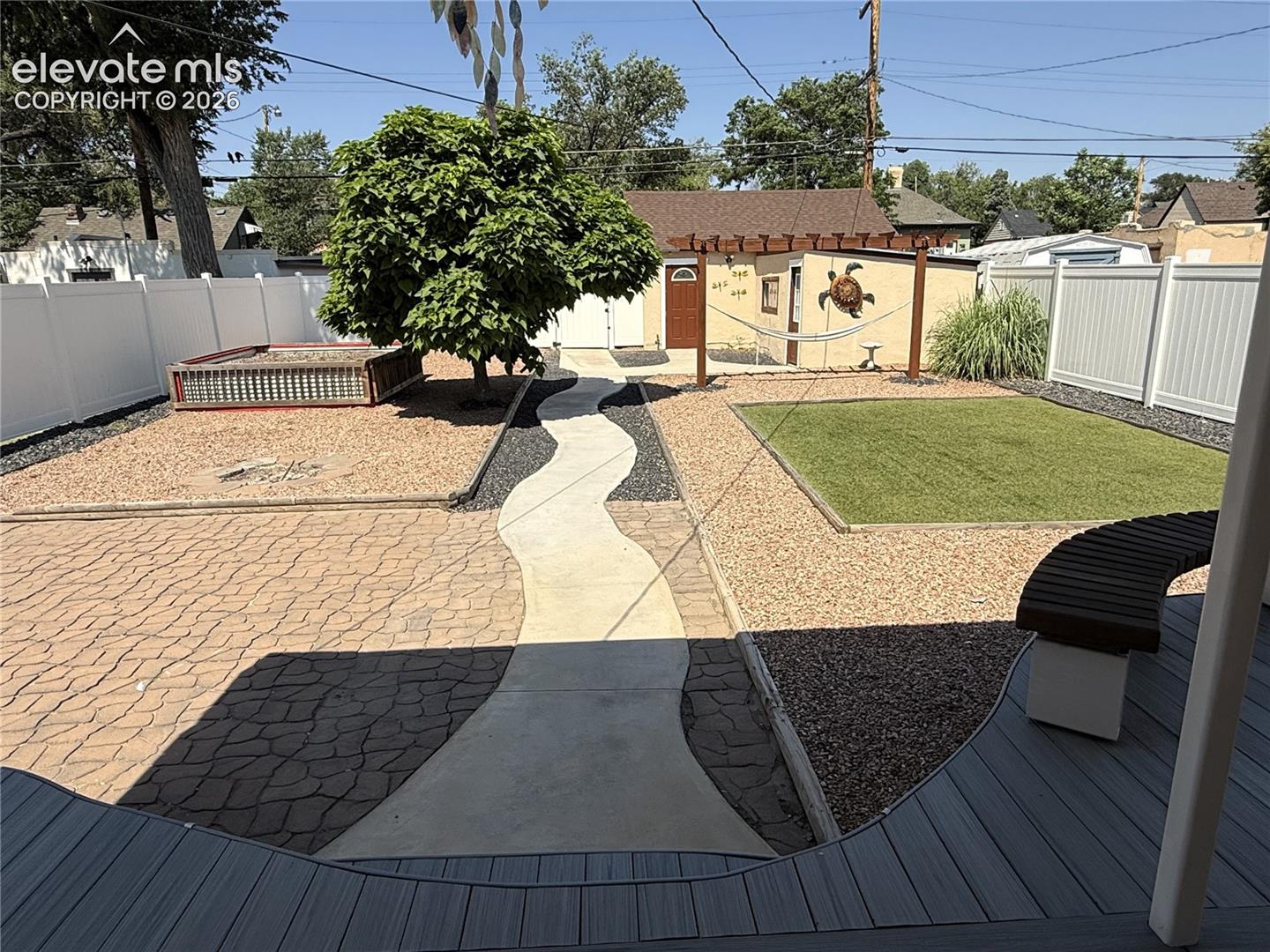 Backyard featuring a composite deck, a curvilinear concrete walkway, stamped hardscaping, low-maintenance gravel beds, and a white vinyl privacy fence