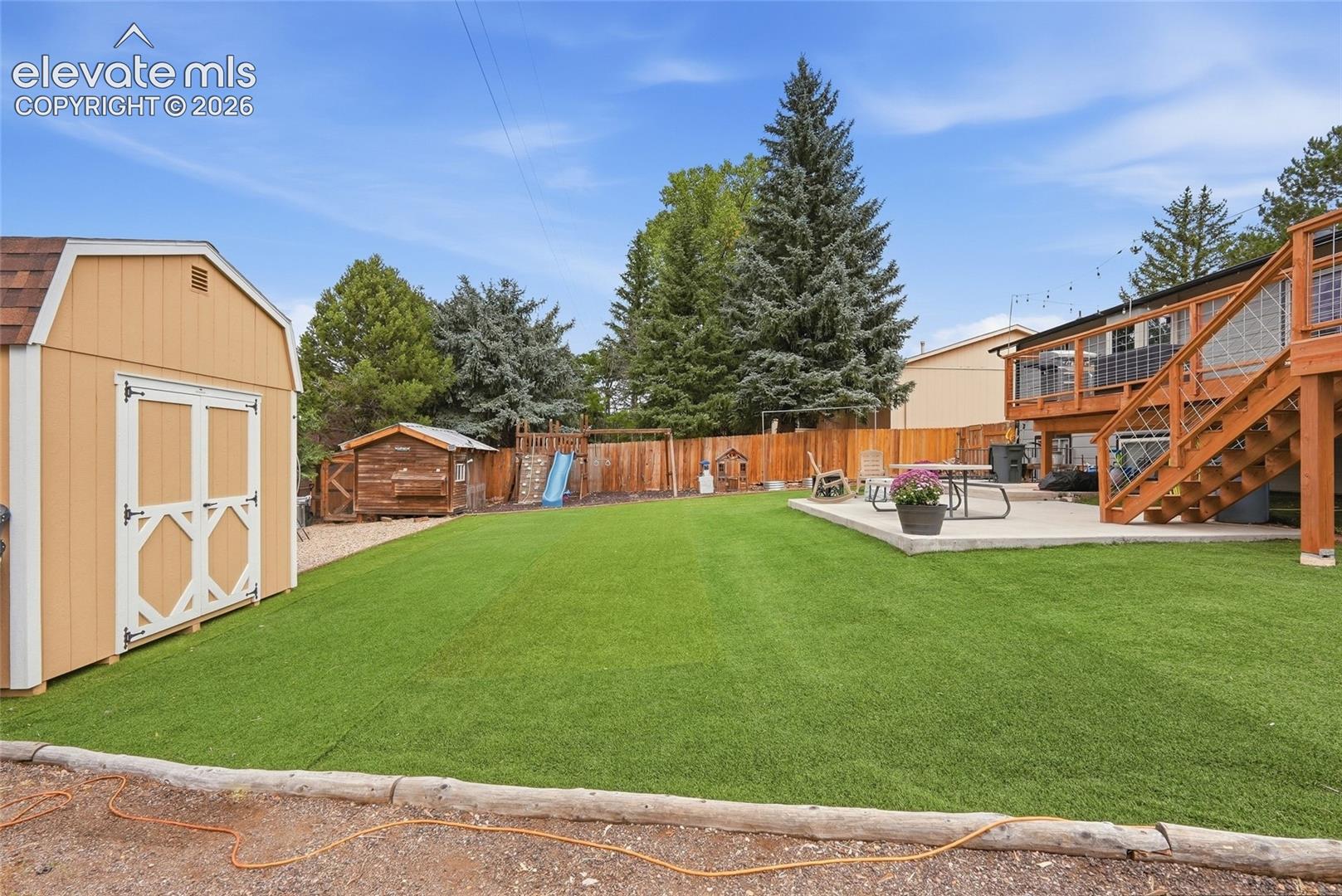 Expansive artificial turf lawn, large concrete patio, multi-level wood deck with stairs, wood privacy fence, and a gambrel-style storage shed