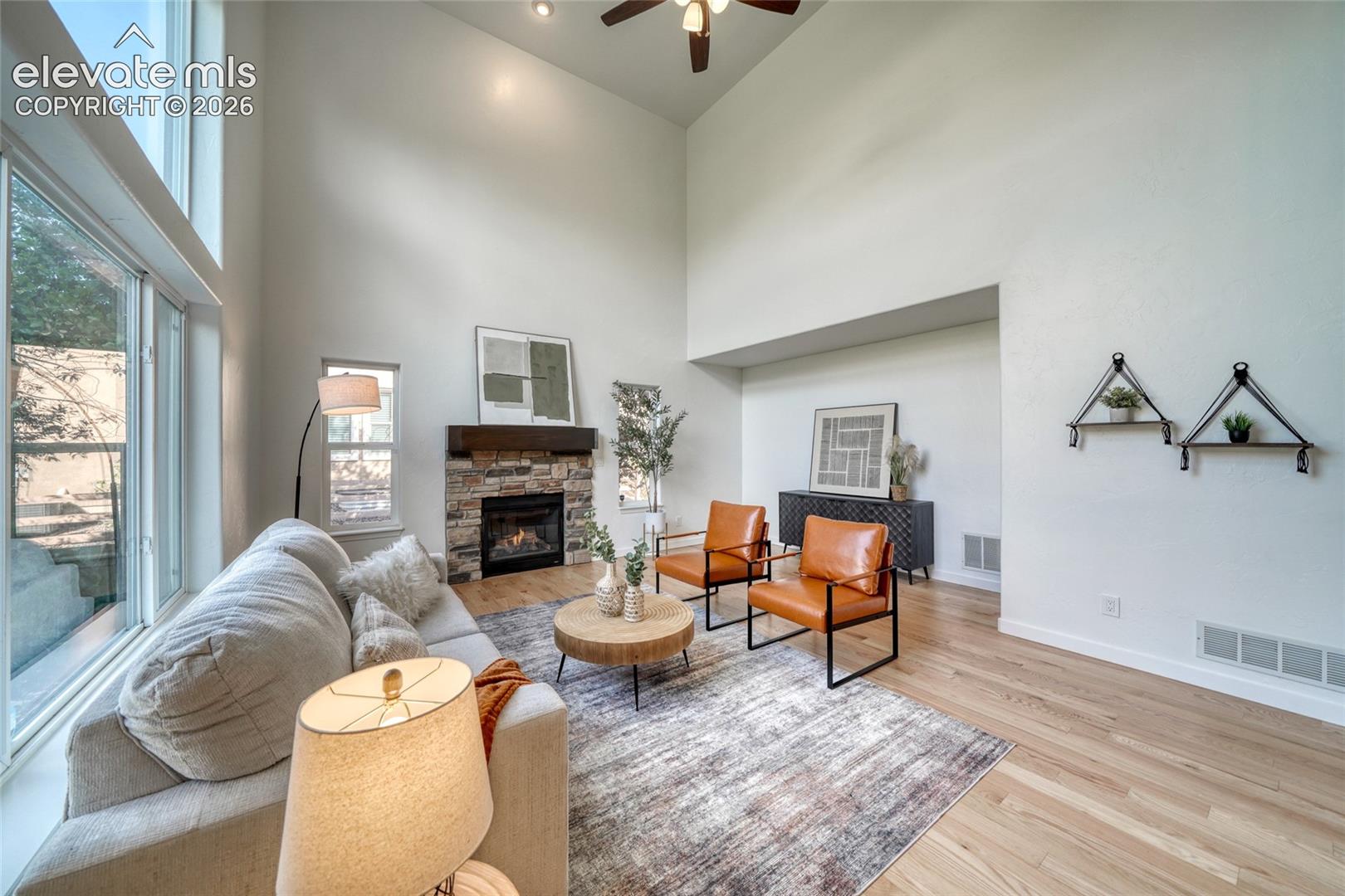 Soaring ceilings with recessed lighting, a stone-faced fireplace with a wood mantle, large windows, and light wood-finish flooring