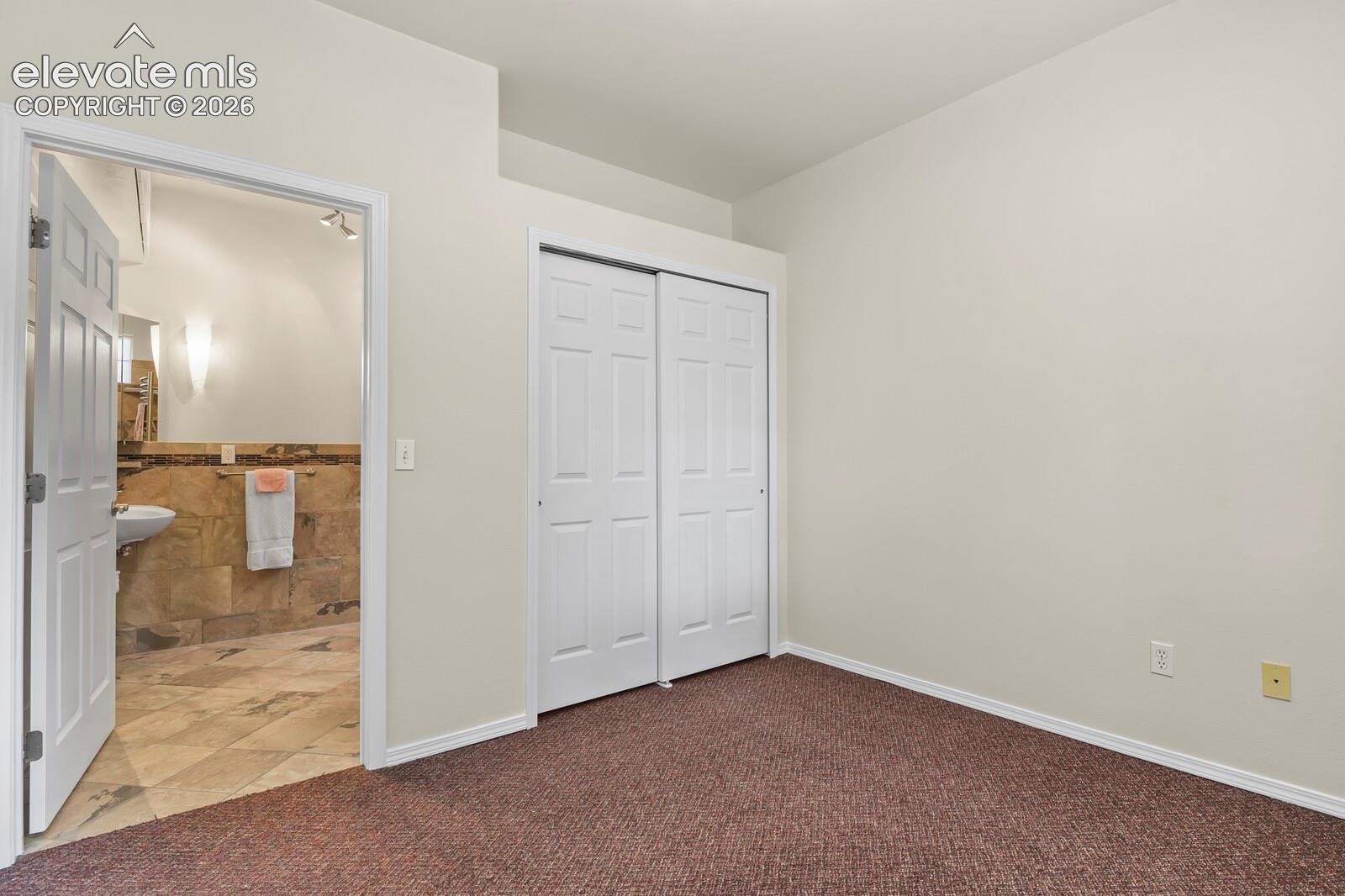 Interior room featuring neutral wall paint, carpeted flooring, and a six-panel sliding closet door, with access to a bathroom finished with stone-tile walls and flooring