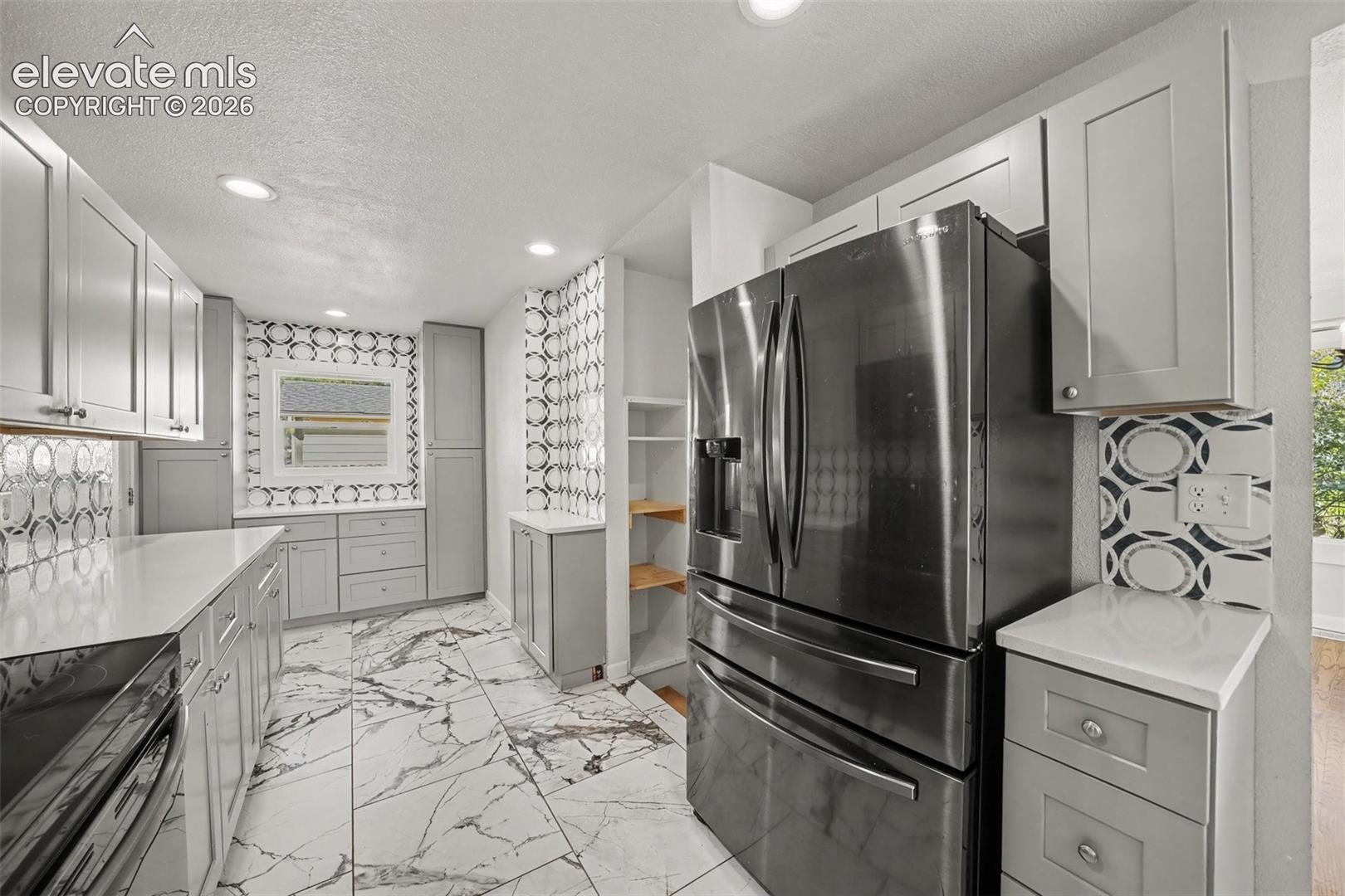 Kitchen featuring grey shaker-style cabinetry, solid-surface countertops, patterned tile backsplash, stainless steel refrigerator, and large-format tile flooring