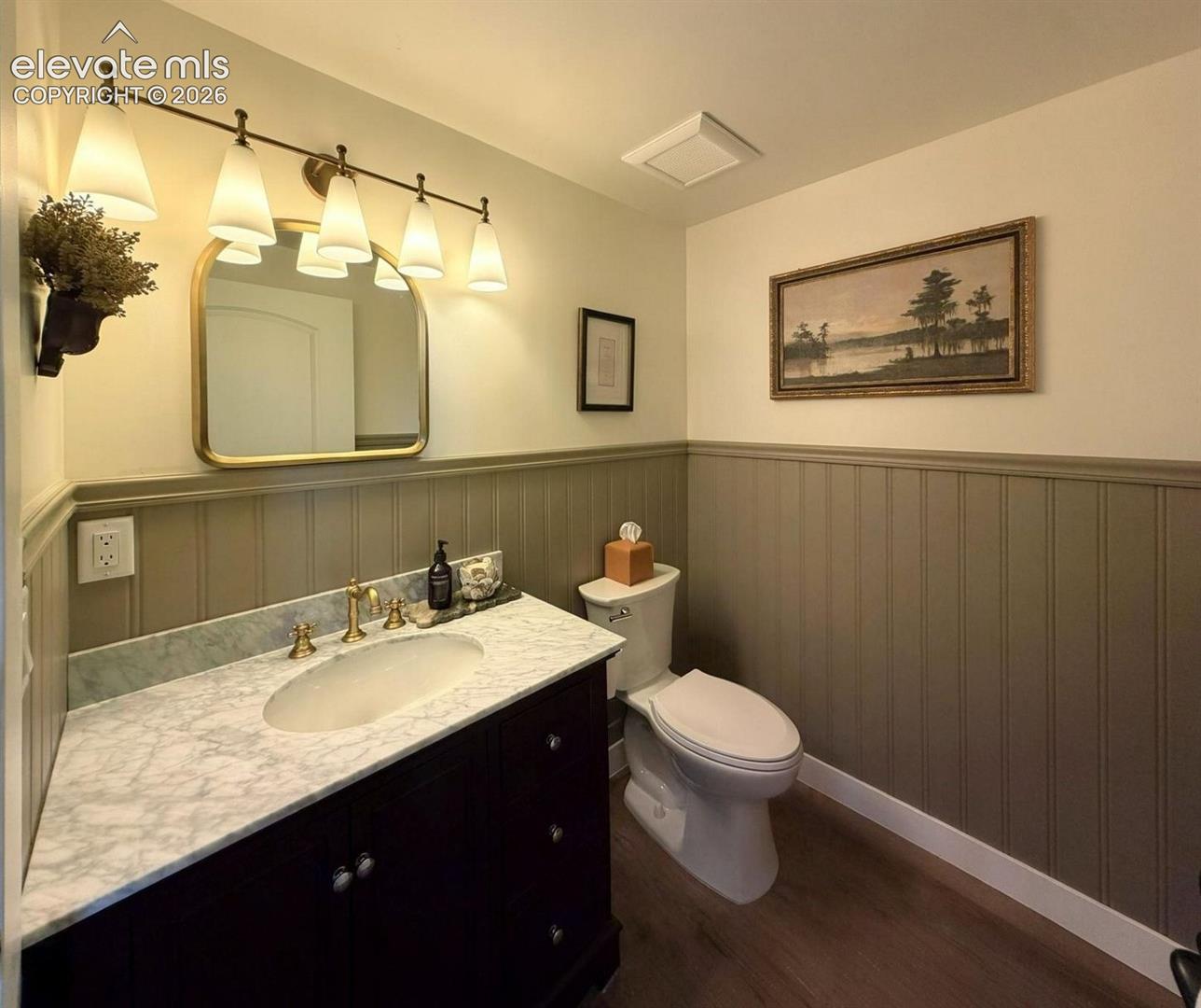 Dark wood vanity with a stone-surface countertop, integrated oval sink, brass-finish faucet, arched wall mirror, and wainscoting