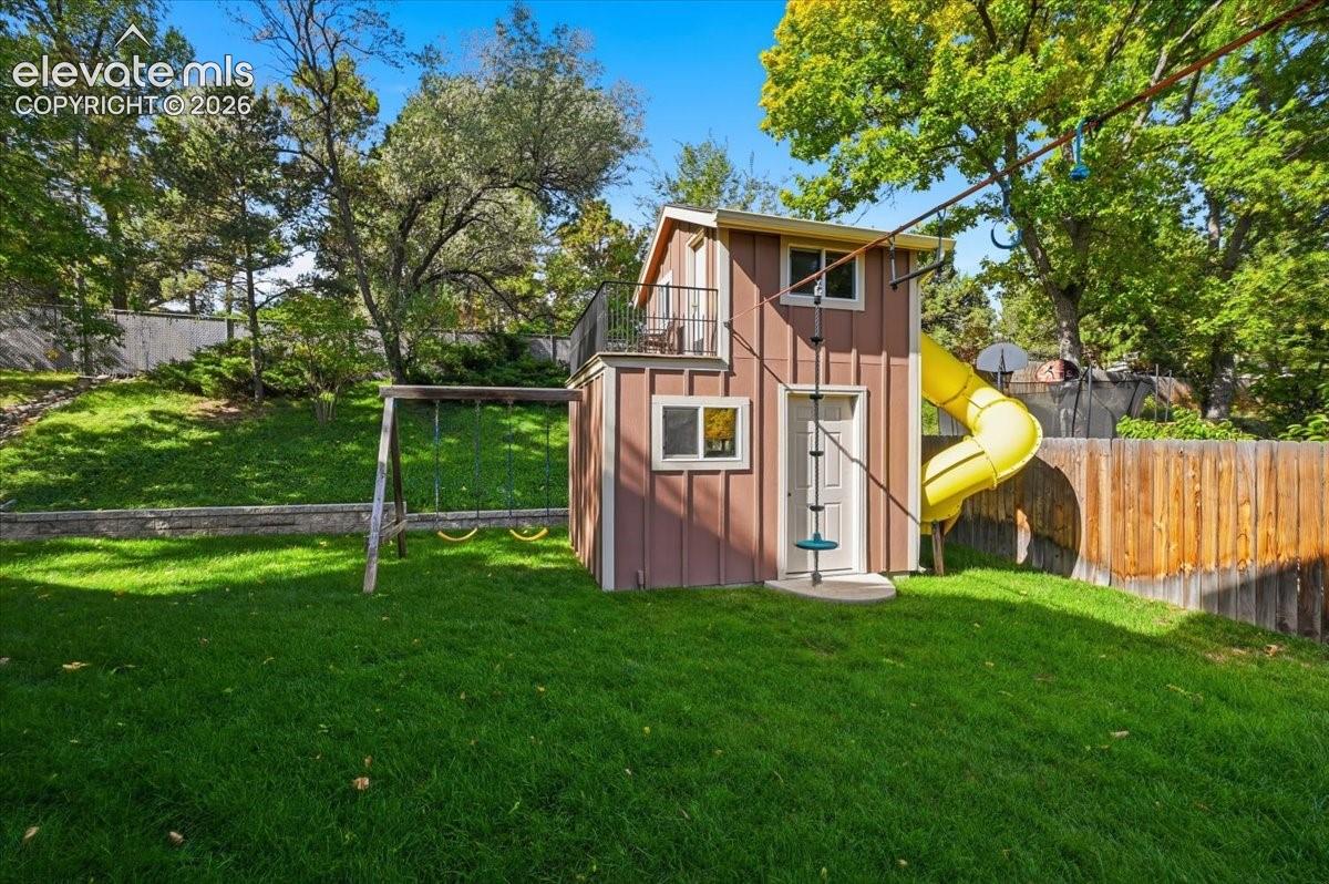 Expansive grassy yard featuring a multi-level playhouse with a spiral slide, a swing set, and a wooden privacy fence