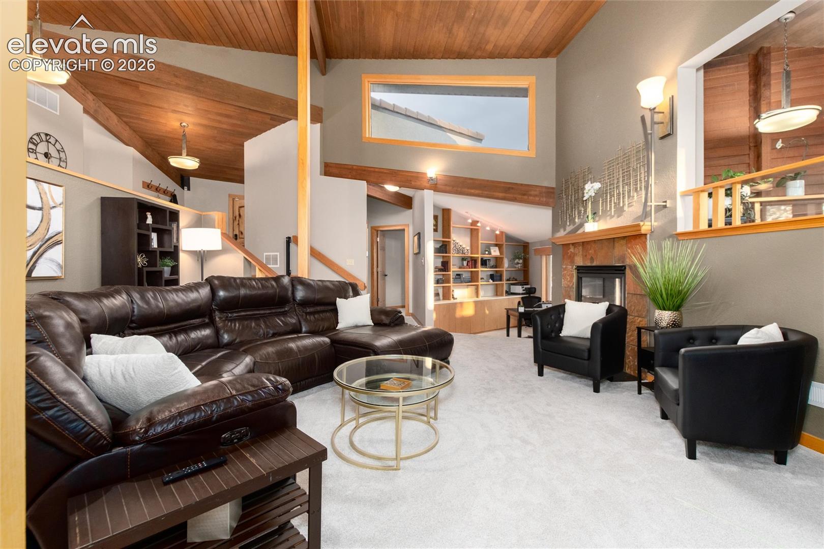Vaulted ceilings with exposed wood beams, light-toned carpeting, a stone fireplace, built-in shelving, and a large upper-level window