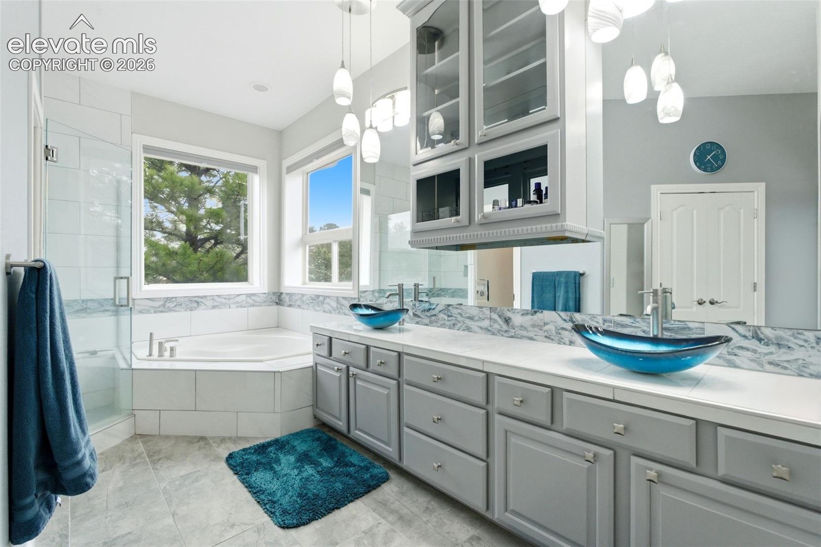 Dual vessel sinks, gray cabinetry, large vanity mirror, soaking tub, and walk-in shower with glass enclosure