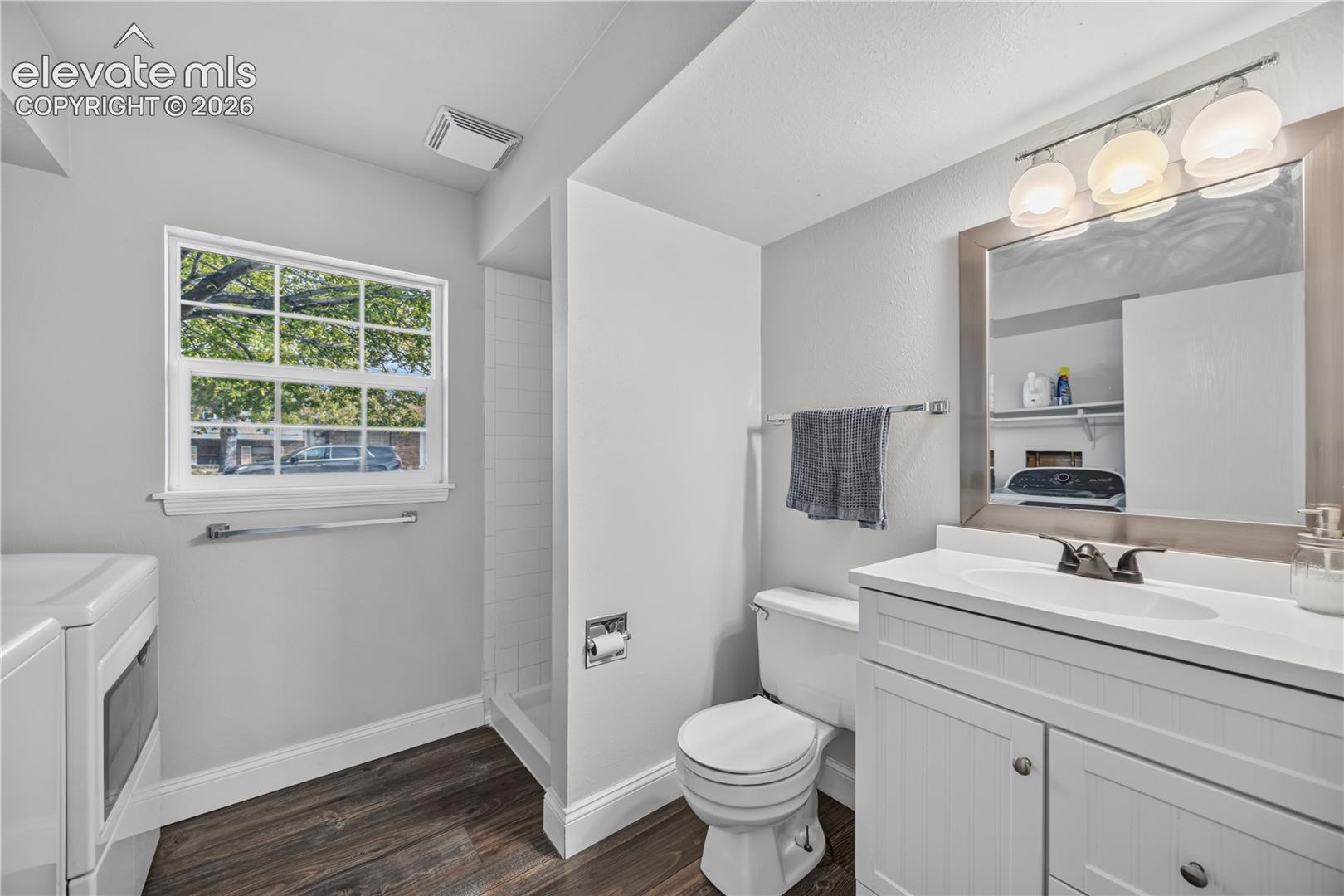 White shaker-style cabinetry with a countertop vanity, dual-handle faucet, integrated wall mirror, and wood-finish flooring