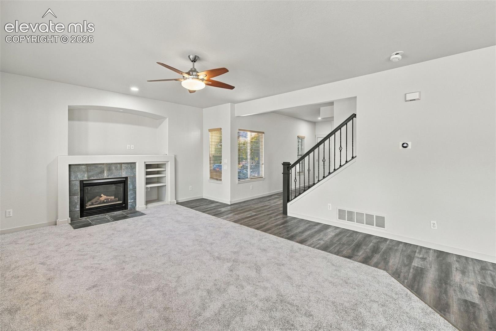 Fireplace with tile surround and built-in shelving, wood-finish flooring, staircase with metal balusters, and ceiling fan with integrated lighting