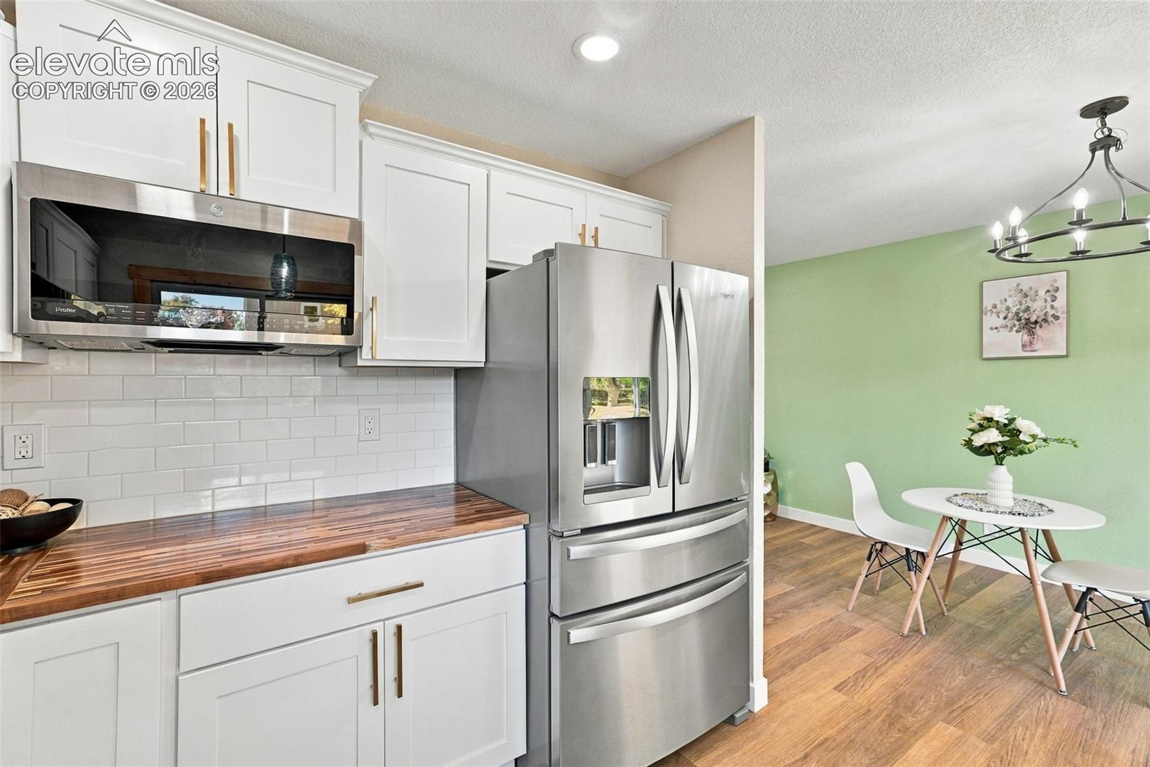 White shaker-style cabinetry with gold-tone hardware, butcher block countertops, subway tile backsplash, stainless steel appliances, and wood-finish flooring