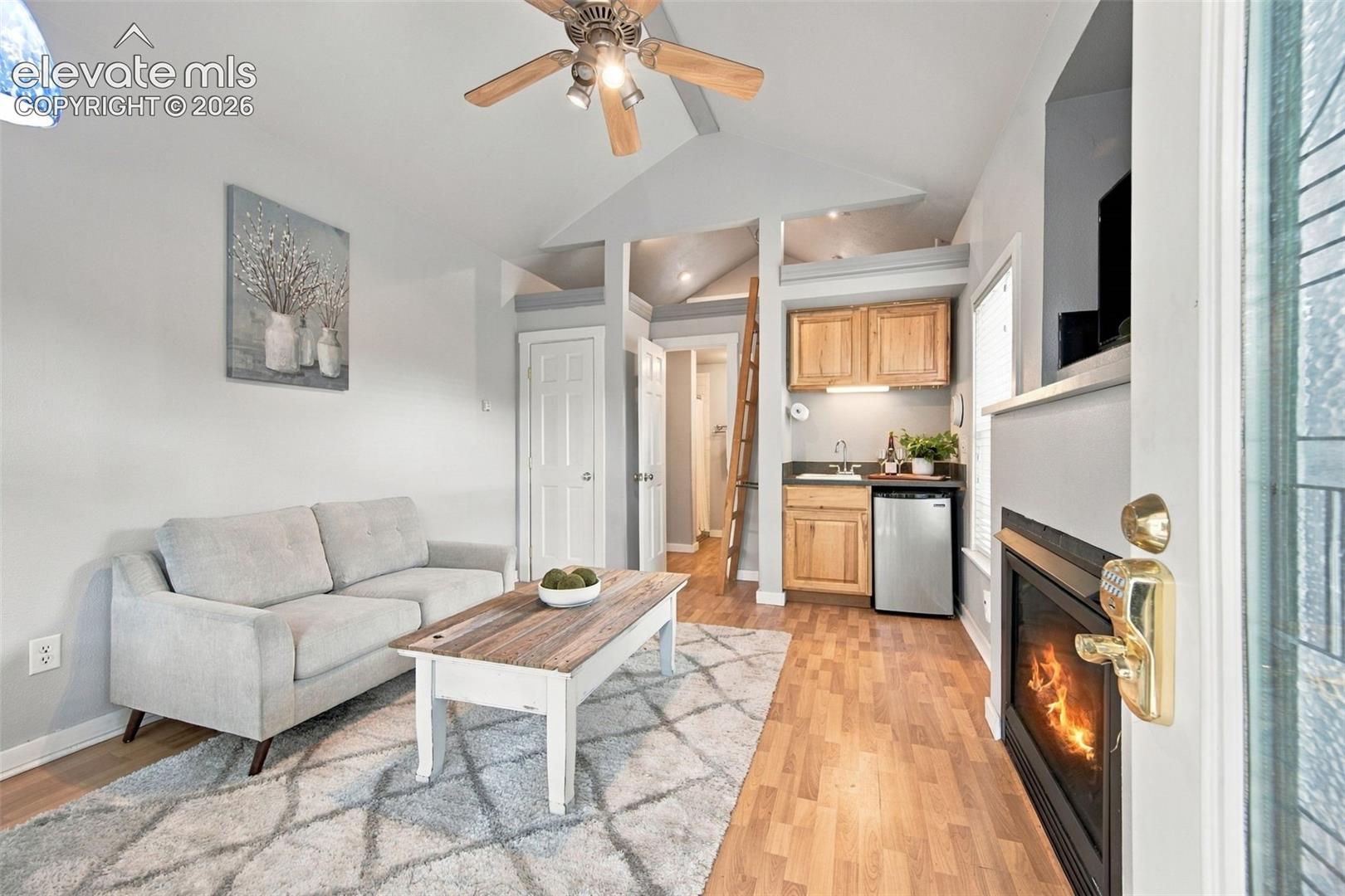 Vaulted ceiling, wood-finish flooring, built-in wet bar with wooden cabinetry, and wall-mounted fireplace