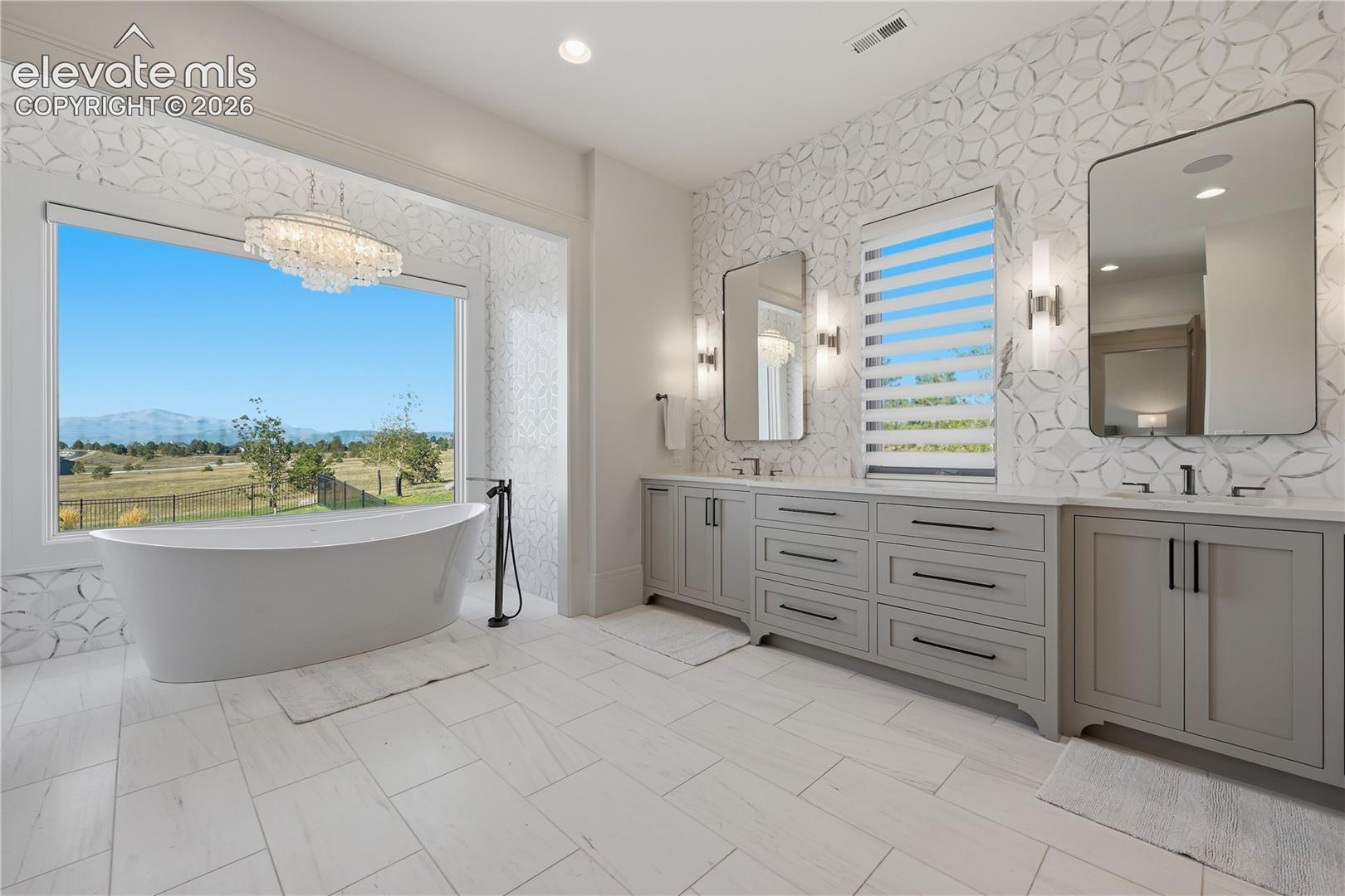 Freestanding soaking tub, dual-sink vanity with matte black hardware, geometric-patterned tile wall, large picture window with mountain backdrop, and light-toned tile flooring
