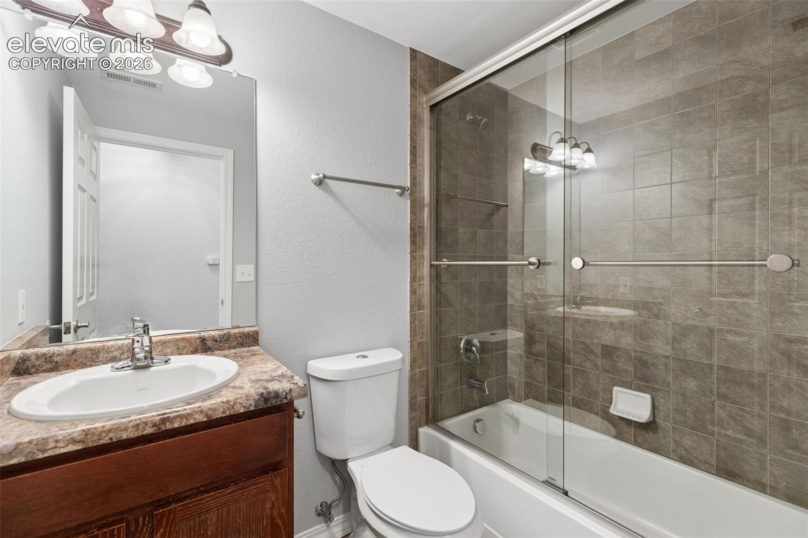 Bathroom featuring a wooden vanity, stone-finish countertop with an integrated oval sink, and a shower-tub combination with tiled surround and glass sliding doors