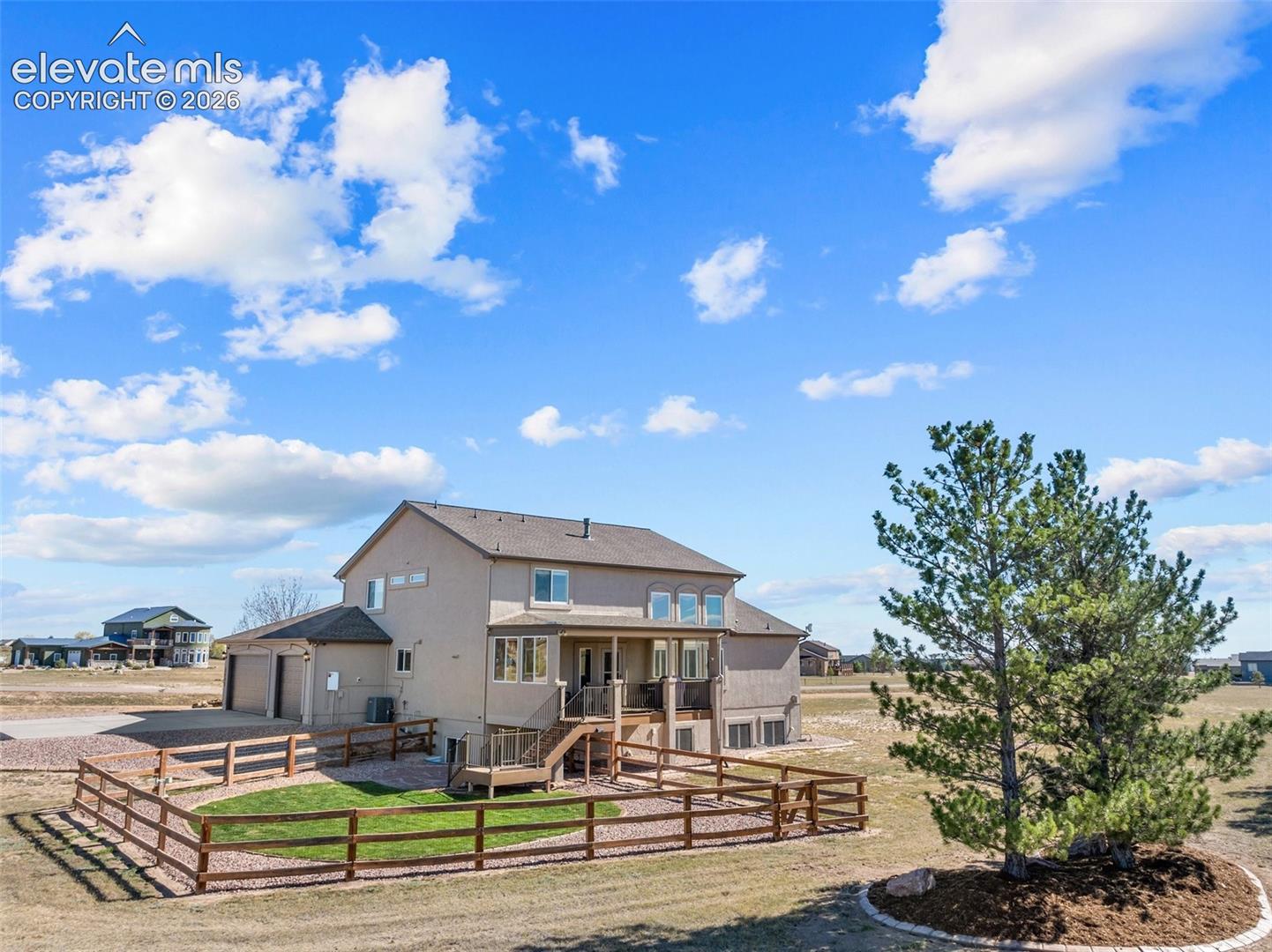 Two-story exterior featuring tan siding, a raised deck with metal railings, a wooden staircase, an attached multi-car garage, and a fenced backyard with landscaped lawn and gravel ground cover