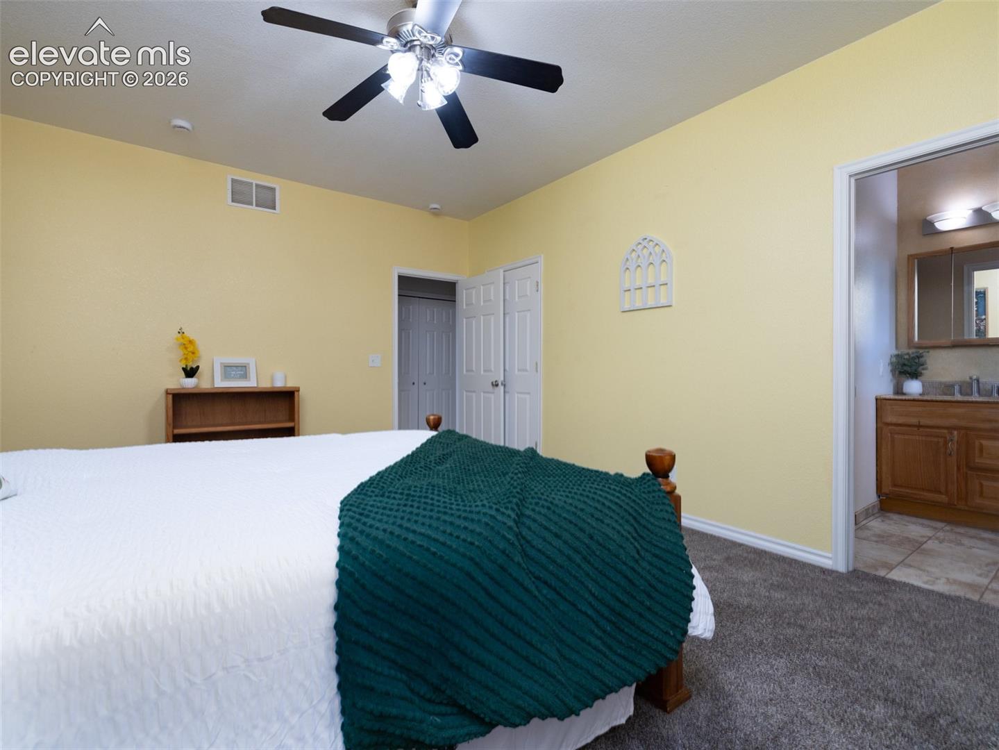 Ceiling fan with lighting, light-toned wall paint, carpet flooring, bi-fold closet doors, and an open doorway leading to a bathroom with tile flooring
