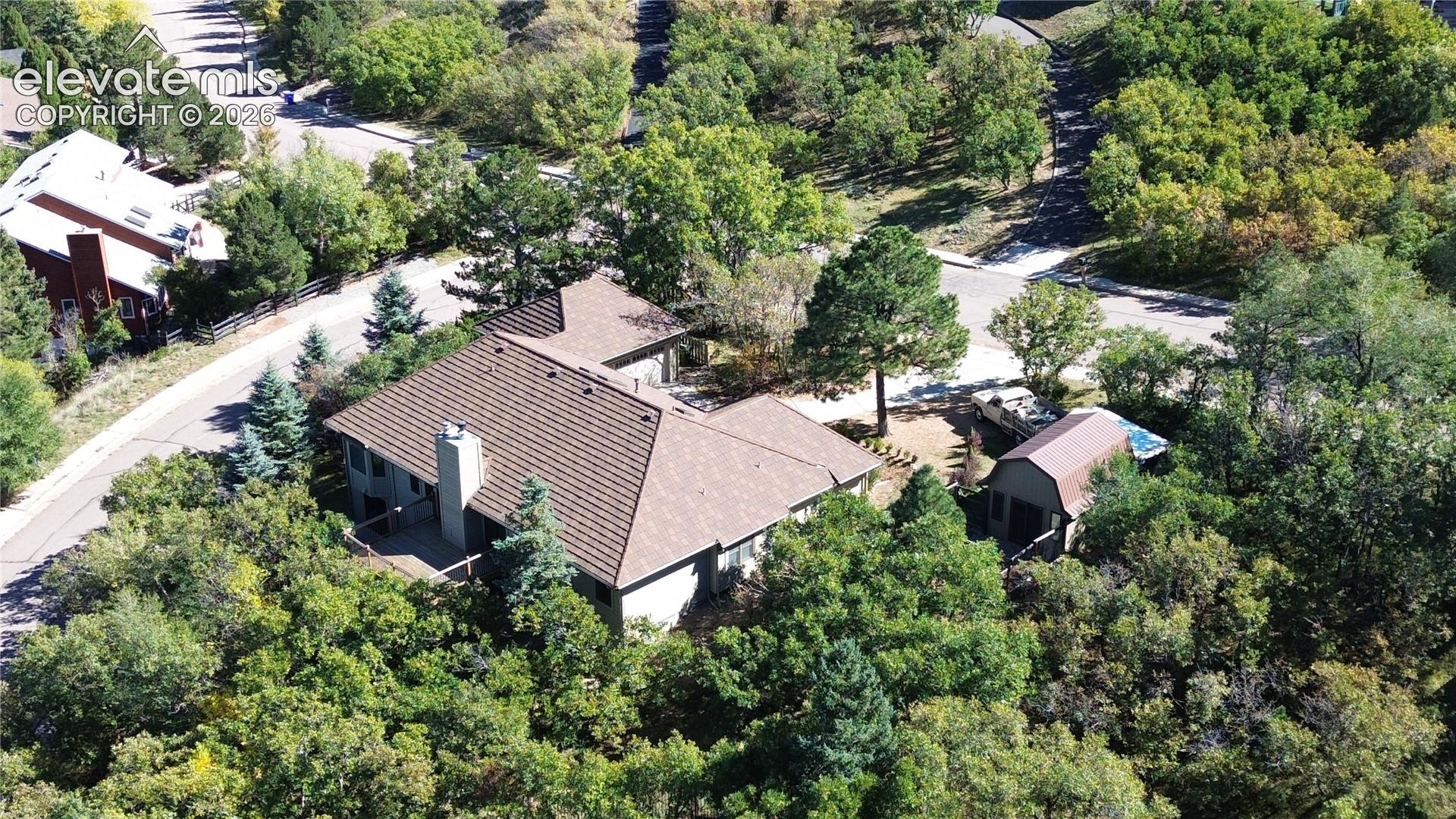 Aerial view featuring a multi-level residence with a shingled roof, attached garage, exterior chimney, and a detached outbuilding, all situated on a lot with extensive tree coverage
