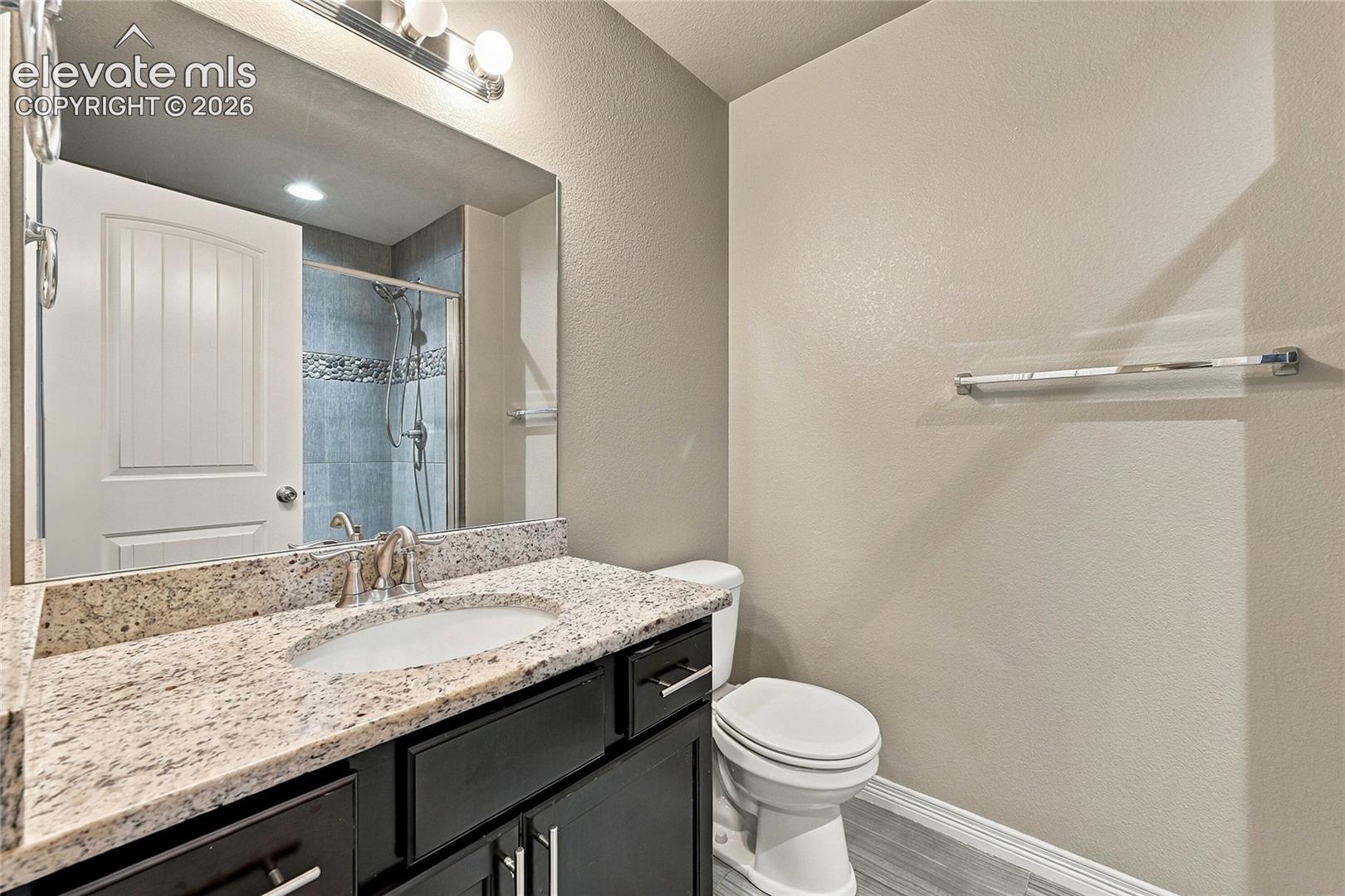 Bathroom featuring a vanity with stone-finish countertop, undermount sink, dark-finish cabinetry, full-width mirror, and a glass-enclosed shower with tiled surround