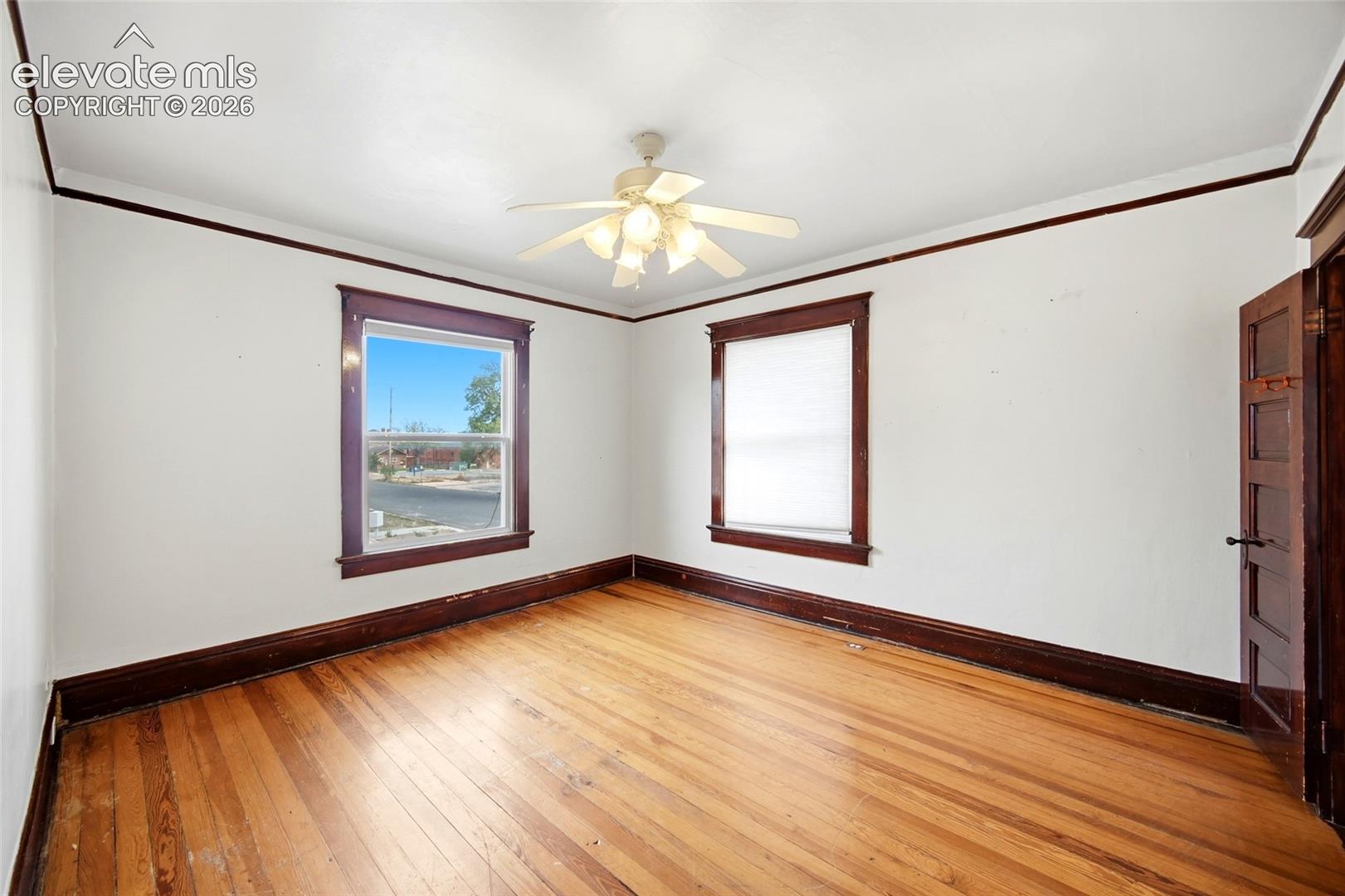 Room interior featuring wood-finish flooring, dark wood-stained window casing, crown molding, a five-panel interior door, and a ceiling-mounted light fixture with a fan