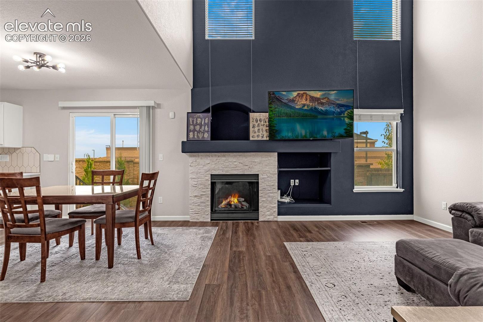 Double-height ceiling, floor-to-ceiling accent wall, fireplace with stone-finish surround, wood-finish flooring, and sliding glass door