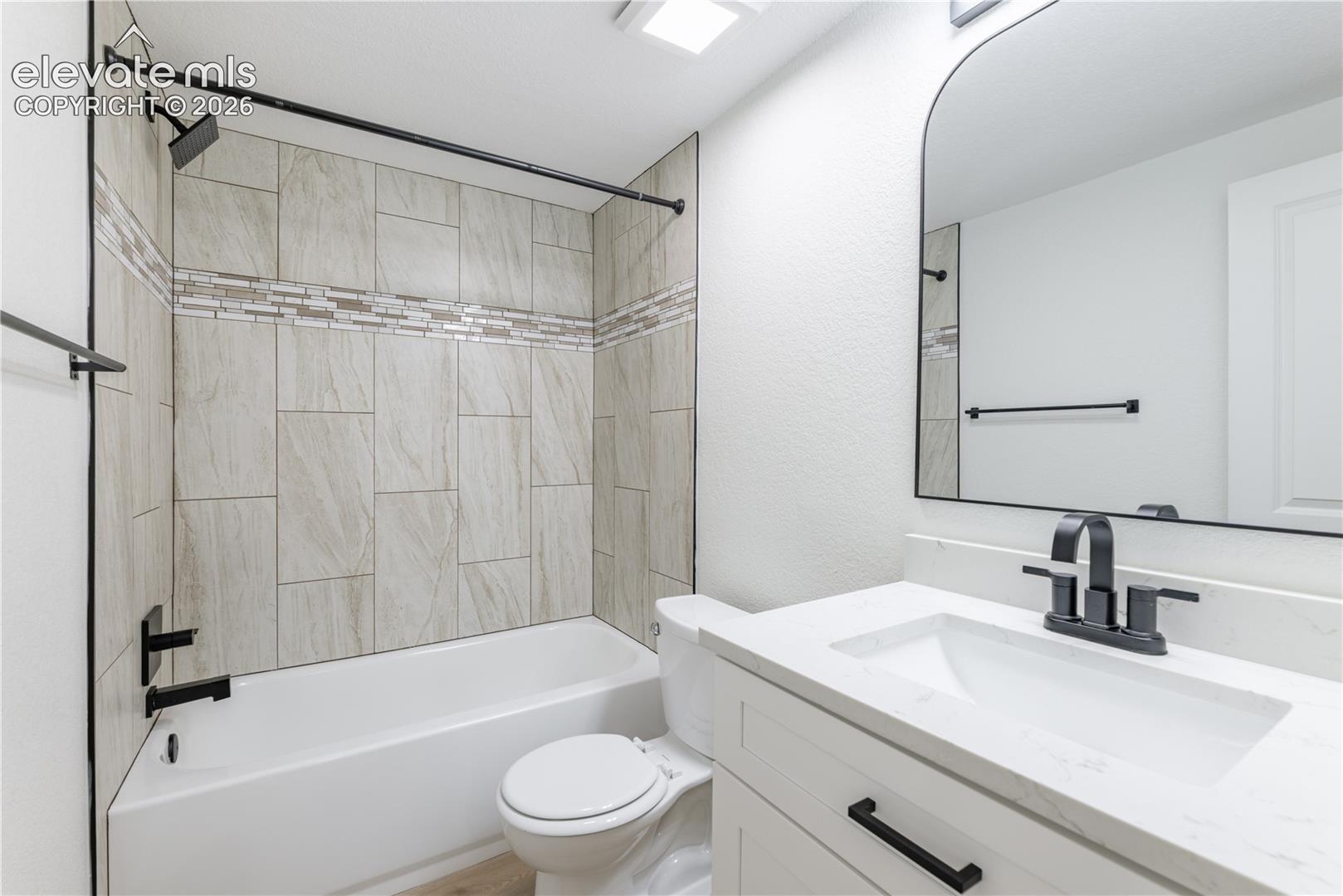 Bathroom featuring a white vanity with a quartz countertop, arched wall mirror, matte black fixtures, and a shower-tub combination with horizontal-set tile surround and decorative glass tile inlay