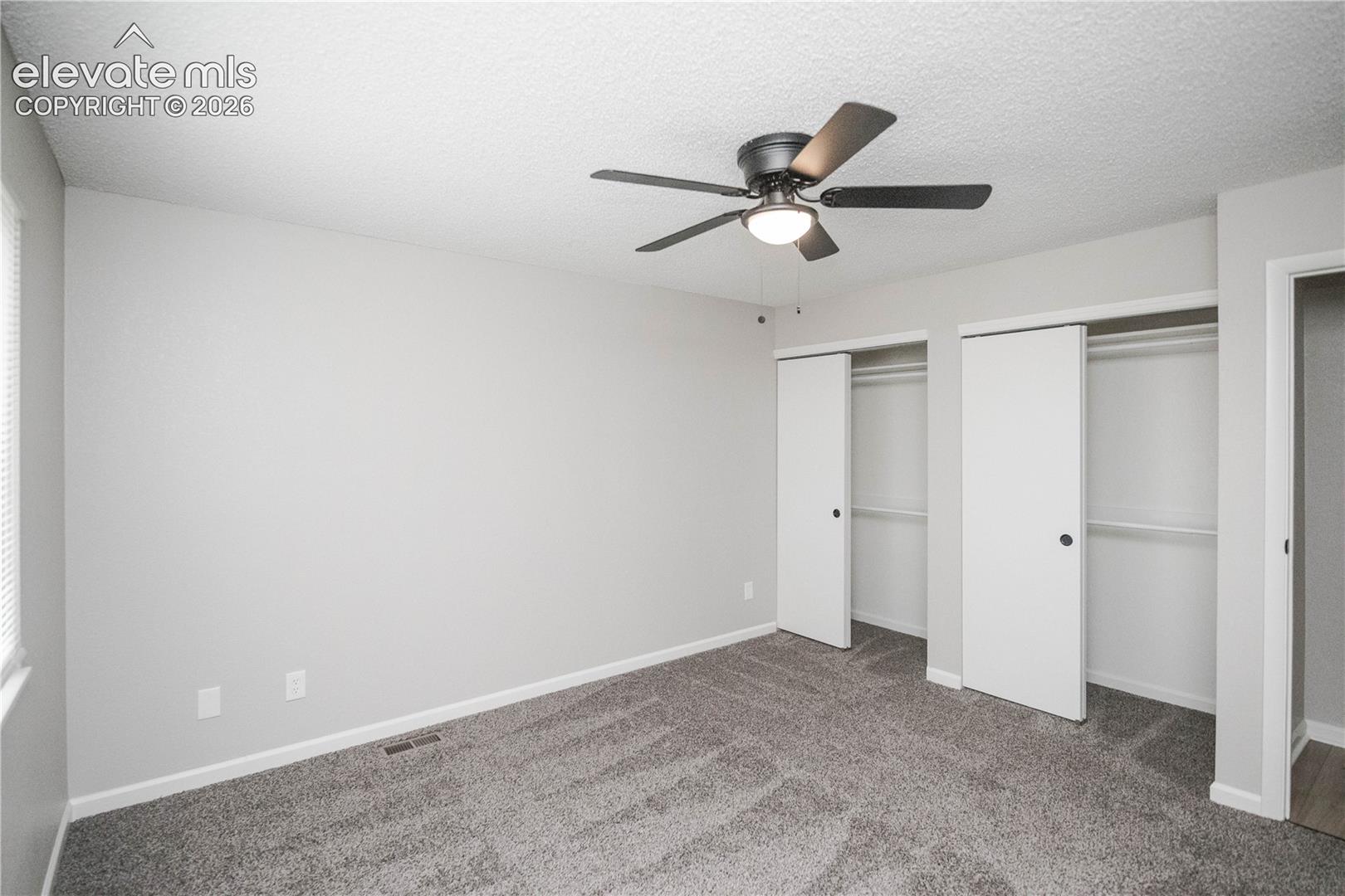 Neutral-toned room featuring textured carpeting, dual closets with swinging doors, and a matte black ceiling fan with an integrated light fixture