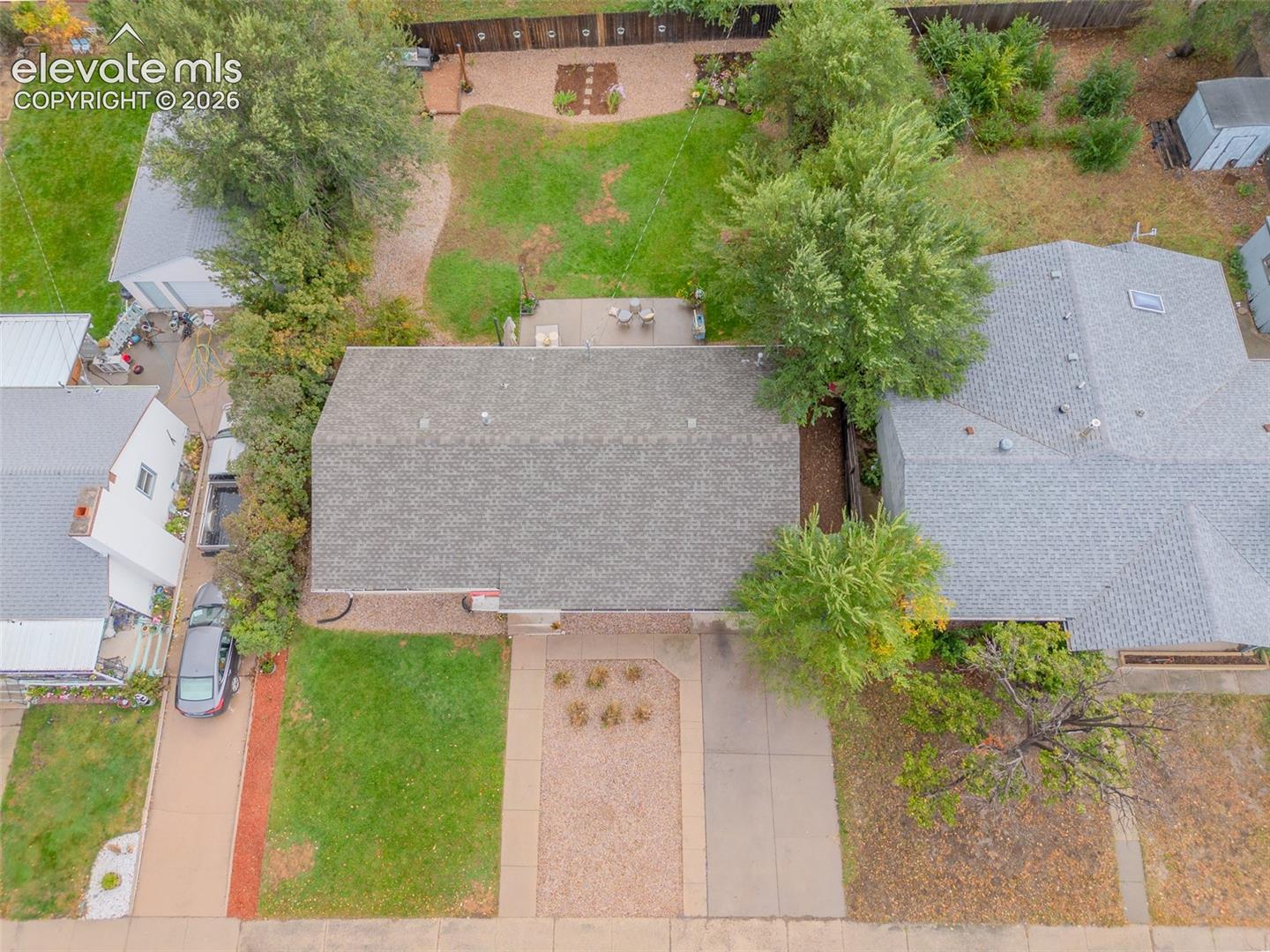 Aerial perspective of property featuring an asphalt shingle roof, a concrete driveway, a rear concrete patio, and a landscaped backyard with mature trees
