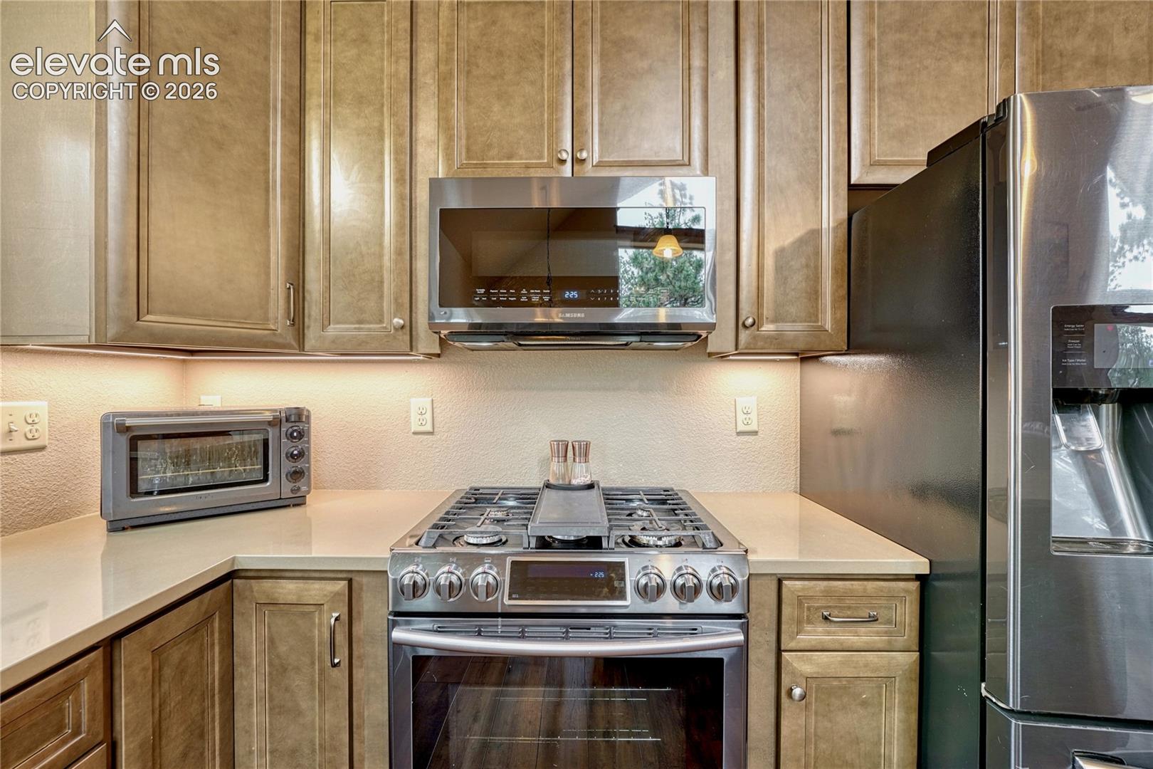 Kitchen featuring stained wood cabinetry, light-toned solid surface countertops, stainless steel gas range, integrated over-the-range microwave, and stainless steel refrigerator
