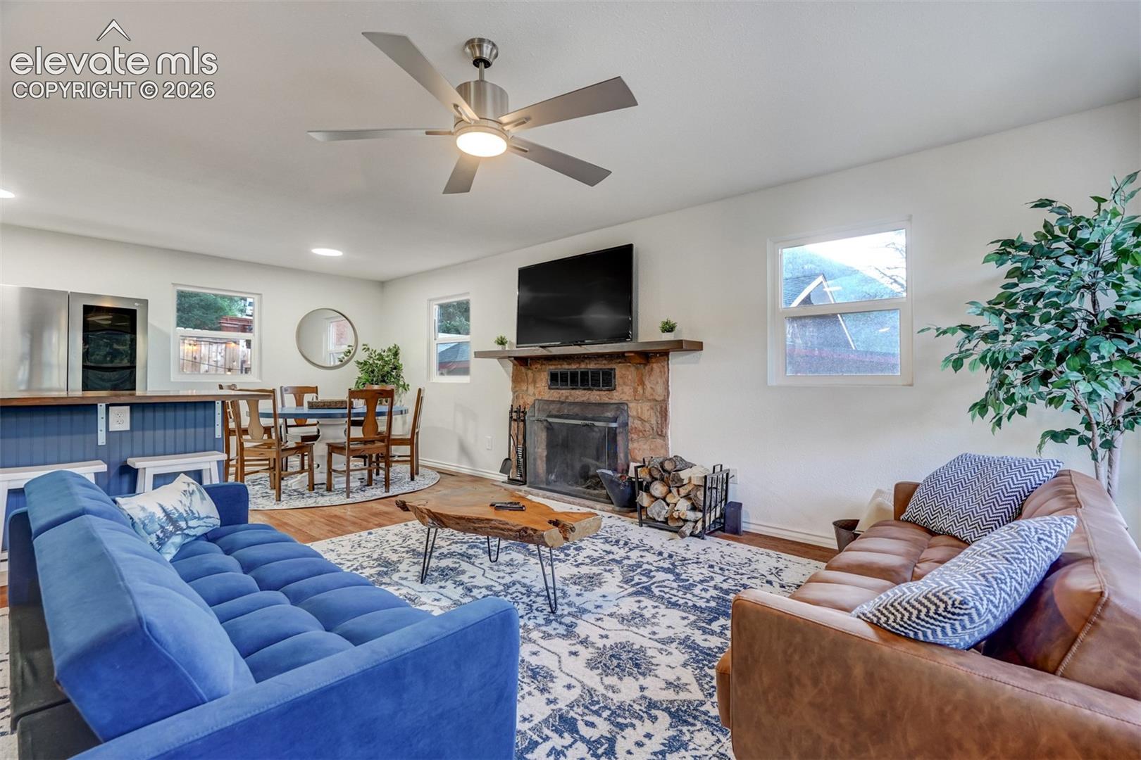 Stone-clad fireplace with a wood mantle, wood-finish flooring, and a modern ceiling fan with integrated lighting
