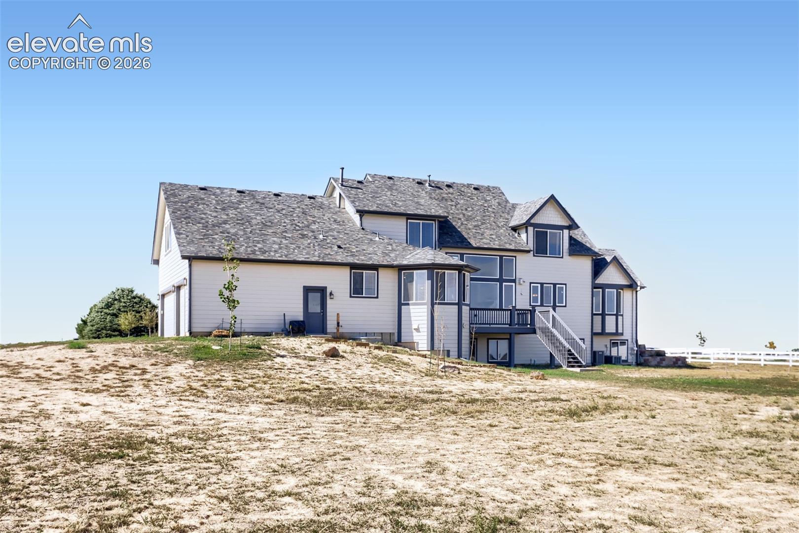 Angled rear view capturing the impressive scope of this multi-level home and its broad, open acreage. Highlighting an elevated back deck, multiple access doors, and expansive usable land with white rail fencing along the perimeter—perfect for livestock, recreation, or future additions.