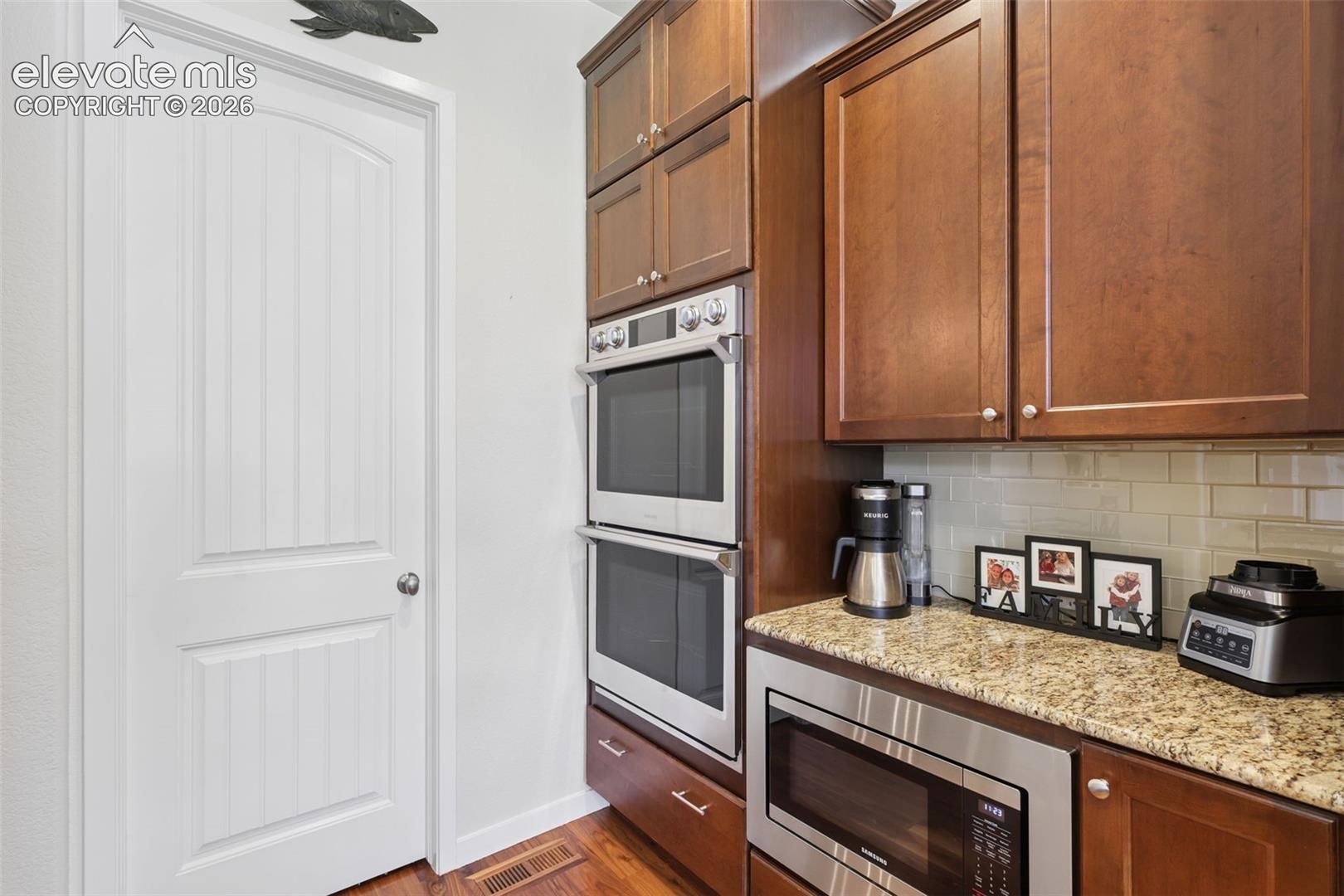 Kitchen featuring wood-finish flooring, stainless steel double wall ovens, a built-in microwave, wood cabinetry, and a granite-finish countertop with a tile backsplash